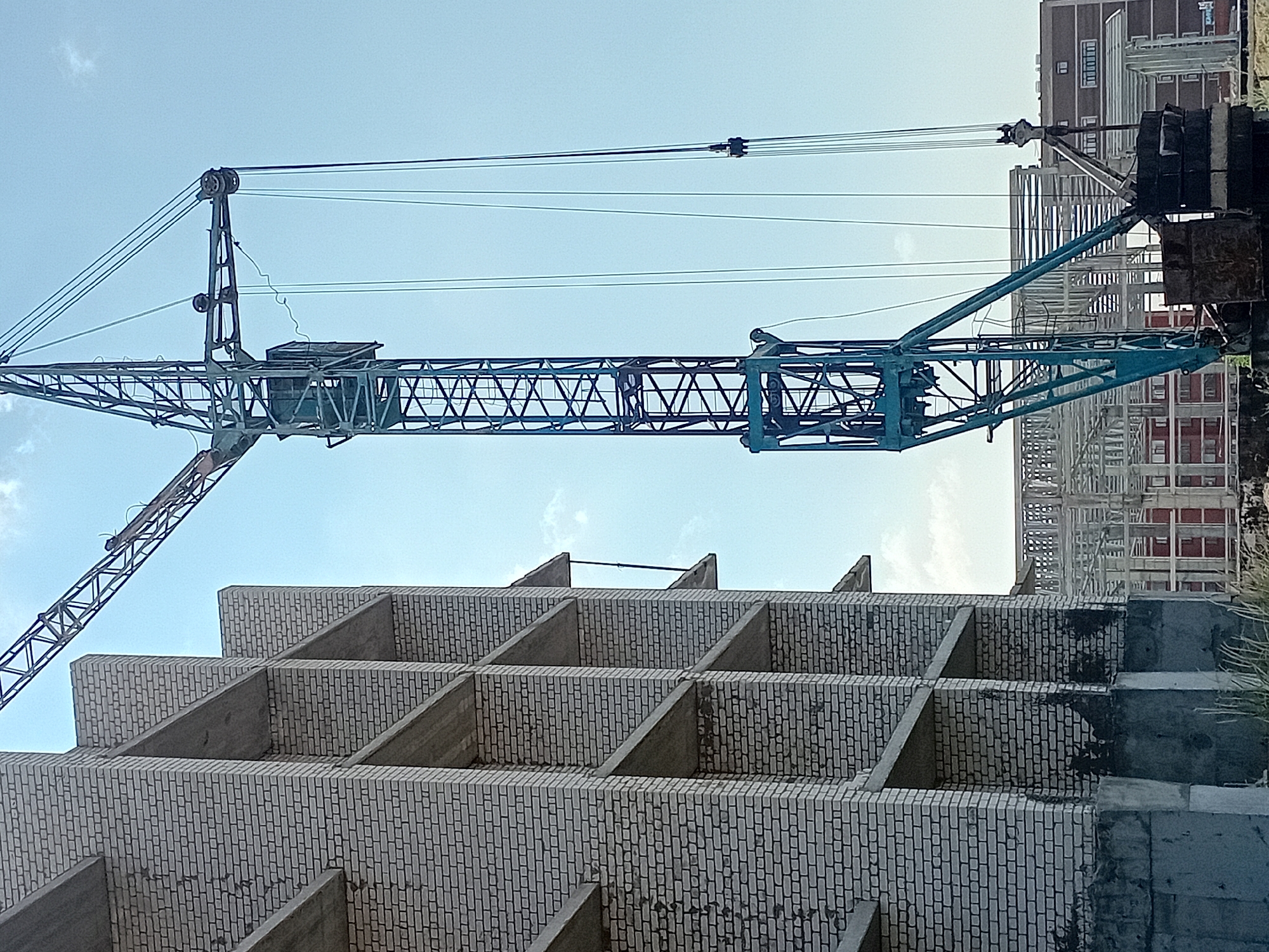 This is a medium-shot, vertical, natural-light photograph taken on a sunny day in the city of Semey, Kazakhstan. The image captures a construction site with an unfinished brick building on the left and a large blue construction crane dominating the center and right portions of the frame. The building is constructed with light-colored bricks and has exposed concrete floors and supports. The crane is an industrial tower crane with a lattice boom, and multiple cables extend from its top. In the background, another partially constructed building with a metal frame is visible, along with a multi-story brick building. The sky is a clear blue with a few faint clouds. There are no people visible in the image. The scene depicts ongoing construction work.