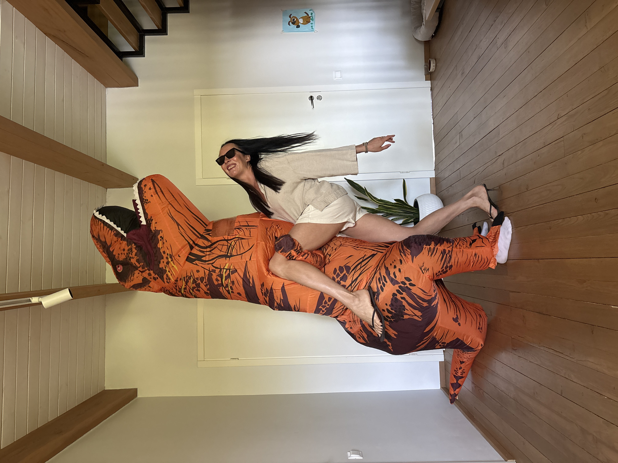 A woman in a cream-colored linen outfit and sunglasses is riding an inflated orange and brown T-Rex costume in a bright, modern interior. The location is a room with light-colored walls and a hardwood floor. The room features wooden beams, a white door, a potted plant, and a framed cartoon dog on the wall. The woman is smiling and has one arm outstretched. The setting may be a residence and is NOT verifiably located in Chaikovsky, Russia.