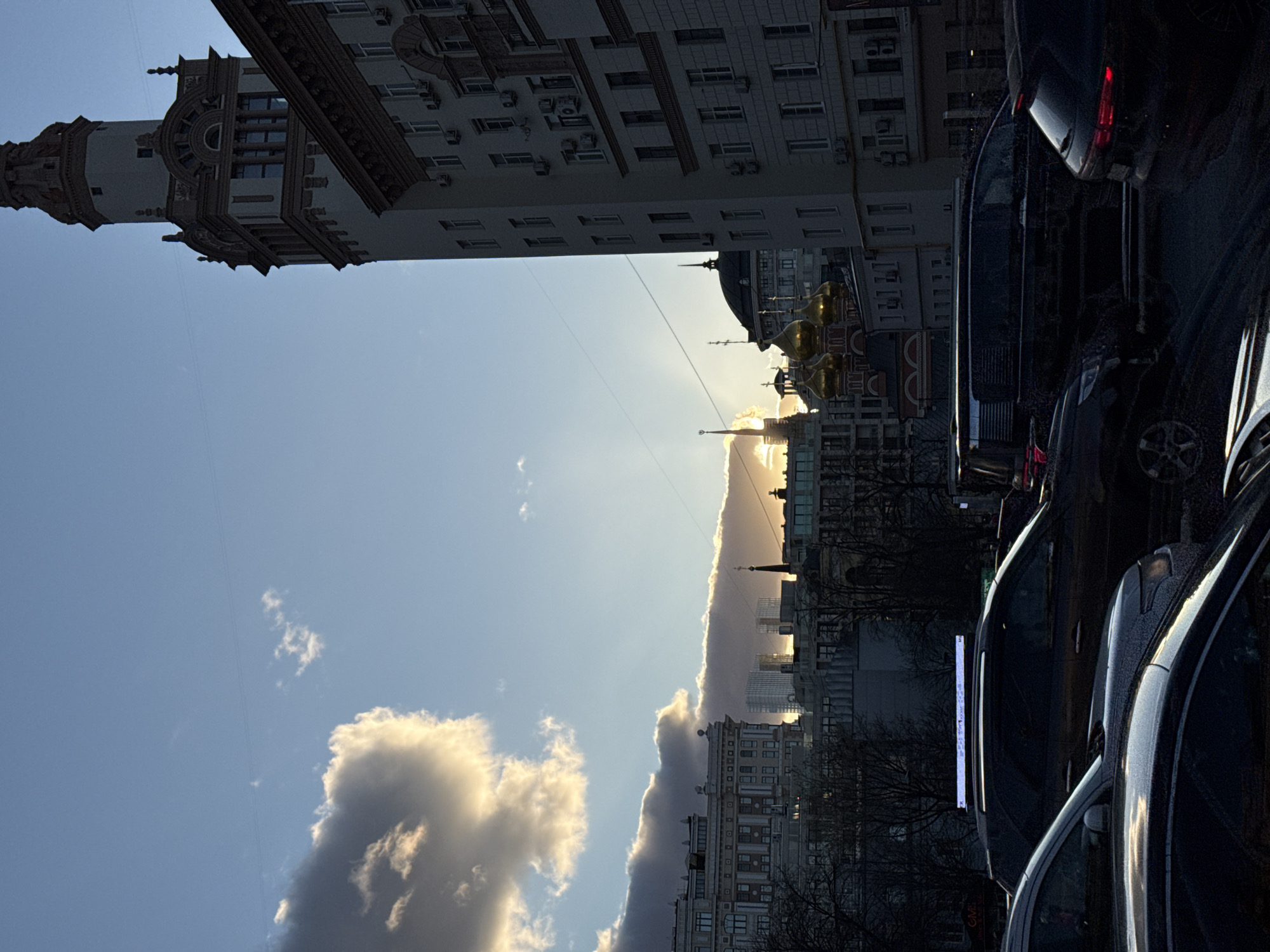 This urban landscape photograph, oriented vertically, captures a dramatic scene during either sunrise or sunset, characterized by striking backlighting. On the left side of the frame, a tall, ornate building with intricate architectural details, possibly in the Stalinist Empire style, rises majestically against the sky. Its complex facade, adorned with cornices and windows, appears partially shadowed.
The central and upper portions of the image are dominated by the sky, which transitions from a clear, soft blue at the top to a warm, golden glow along the horizon. A prominent, elongated cloud formation stretches horizontally across the mid-ground, brilliantly backlit by the low sun, creating a vivid line of golden light and a dramatic silhouette. Below this illuminated cloud, the distant cityscape reveals various buildings, including what appear to be modern high-rises and structures with distinctive golden domes, hinting at a church or cathedral, all subtly touched by the golden light.
To the right, a line of several parked cars, predominantly dark sedans, occupies the foreground and mid-ground. Their surfaces reflect fragments of the sky's light, creating subtle highlights against their shadowy forms. Bare tree branches are visible in the lower right corner, suggesting a late autumn or winter season.
The scene is devoid of overt human activity, conveying a sense of serene grandeur. The architectural styles, particularly the ornate building and the golden domes, are strong location cues for Moscow, Russia, as specified. There is no visible text in the image. The dynamic interplay of light and shadow, combined with the distinctive urban architecture, creates a captivating and atmospheric view of the city.