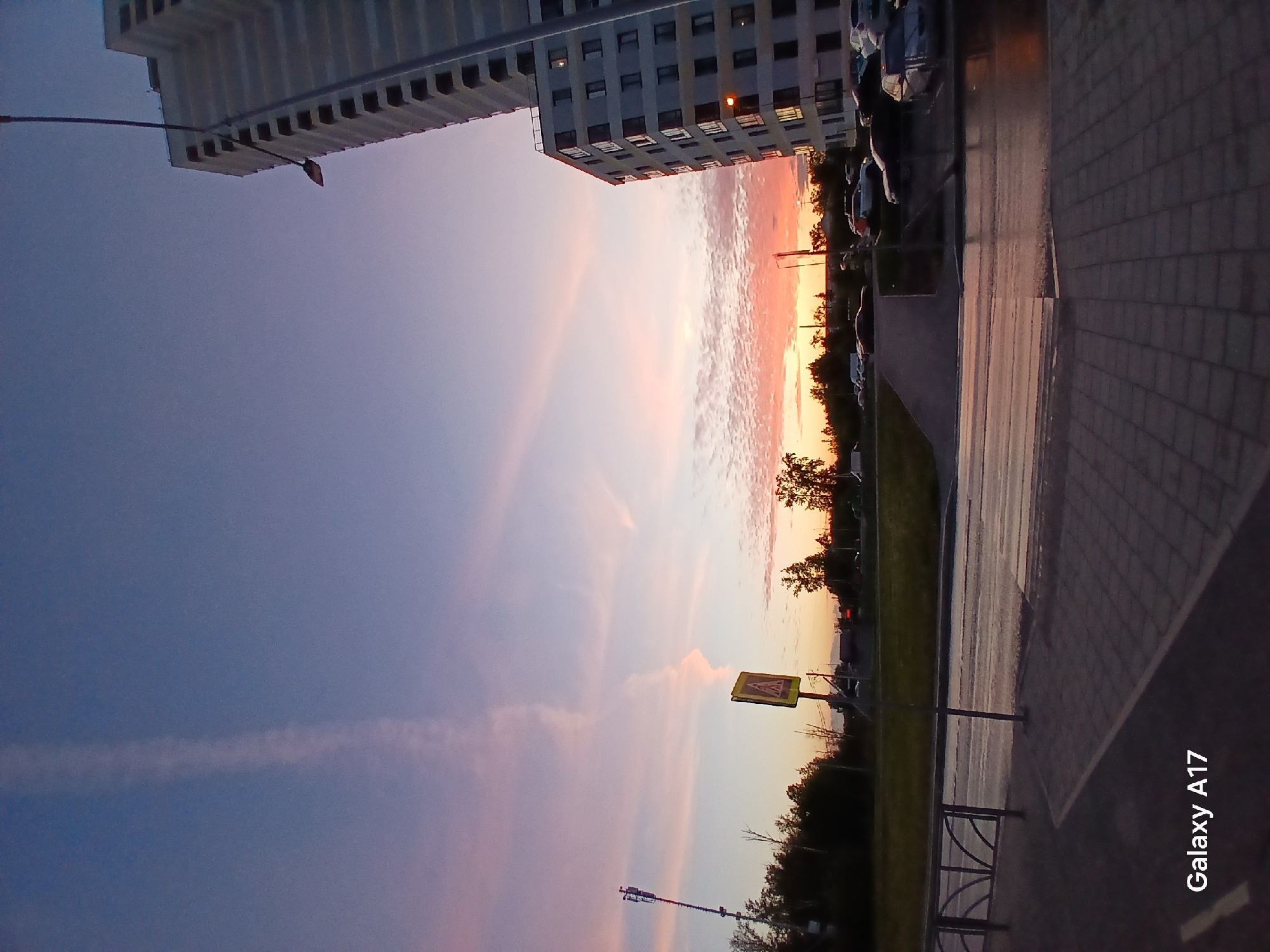The image is of a sunset in the city of Yekaterinburg, Russia, taken from a low angle. The sky displays a gradient of colors from deep blue to soft pink and orange near the horizon. A white contrail stretches across the upper left sky. Buildings are visible in the upper portion of the frame. A taller white building is visible at the top left with horizontal architectural details. Another mid-rise building can be seen in the top center, with many windows. A row of parked cars lines the street to the right. Trees and vegetation are situated along the mid-ground, with a yellow pedestrian crossing sign also visible. The foreground is dominated by a gray tiled walkway and a low metal fence. A "Galaxy A17" watermark is visible at the bottom right. The light of the setting sun reflects off the wet surfaces of the street.