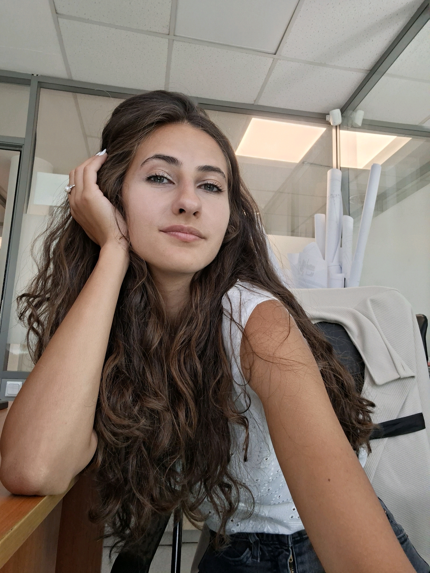 A young woman with long, dark, curly hair and fair skin is captured in a medium shot, resting her head on her hand. Her left arm is propped on a wooden surface, likely a desk, with her elbow forming a curve. She is wearing a white top with intricate eyelet detailing and dark-colored pants, possibly jeans. A light-colored jacket or sweater is draped over the back of a chair behind her. Her gaze is directed slightly upwards and to the left, with a neutral expression. A ring with a pearl is visible on her left hand. In the background, there is office-like partitioning with glass panels, a suspended ceiling with recessed lighting, and several rolled-up documents, suggesting an indoor professional environment. The overall lighting is bright, illuminating the scene clearly.