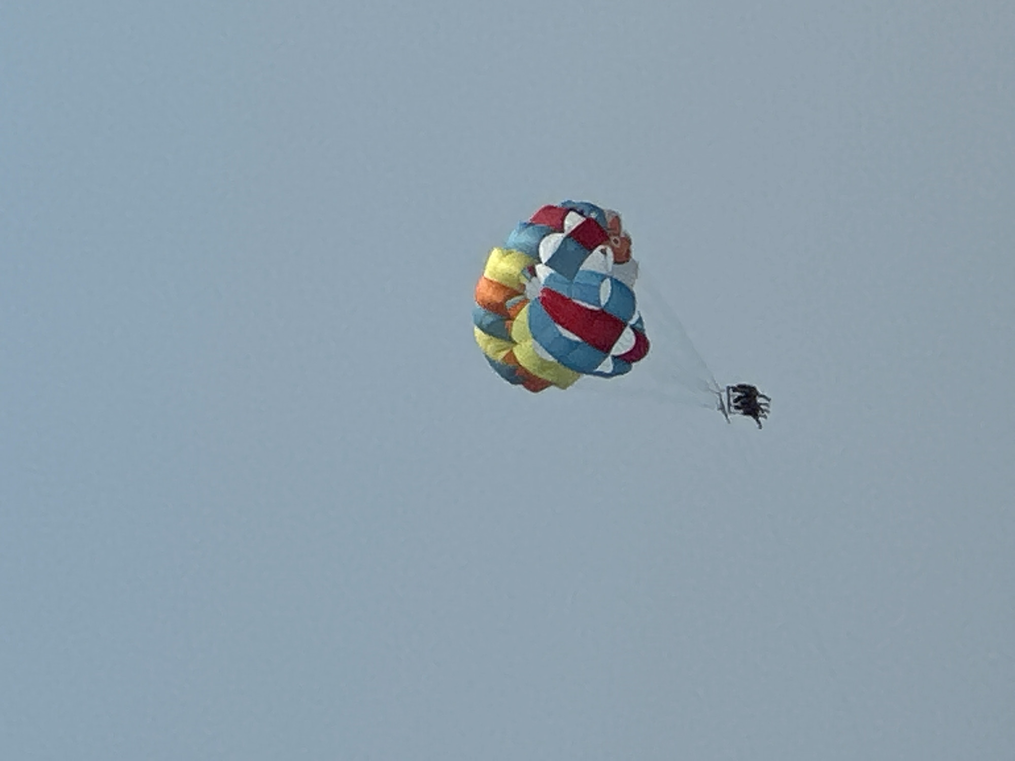 The image shows a parasailer in mid-air against a clear, pale blue sky. The parasail is colorful, with panels of red, white, blue, yellow, and orange. It is inflated and appears to be angled slightly to the right, suggesting it is actively being used for parasailing.
Below the parasail, a small, dark object is visible, which appears to be a harness or seat where the person is suspended. Two figures, likely people, are seated in the harness. Their dark clothing makes them difficult to discern clearly against the sky.
The scene suggests a recreational activity taking place on a bright, clear day, likely during daylight hours. There are no discernible location cues other than the provided context of Boreti, Montenegro, and no visible text. The overall impression is one of freedom and adventure as the individual is lifted high into the sky by the wind-powered parasail.