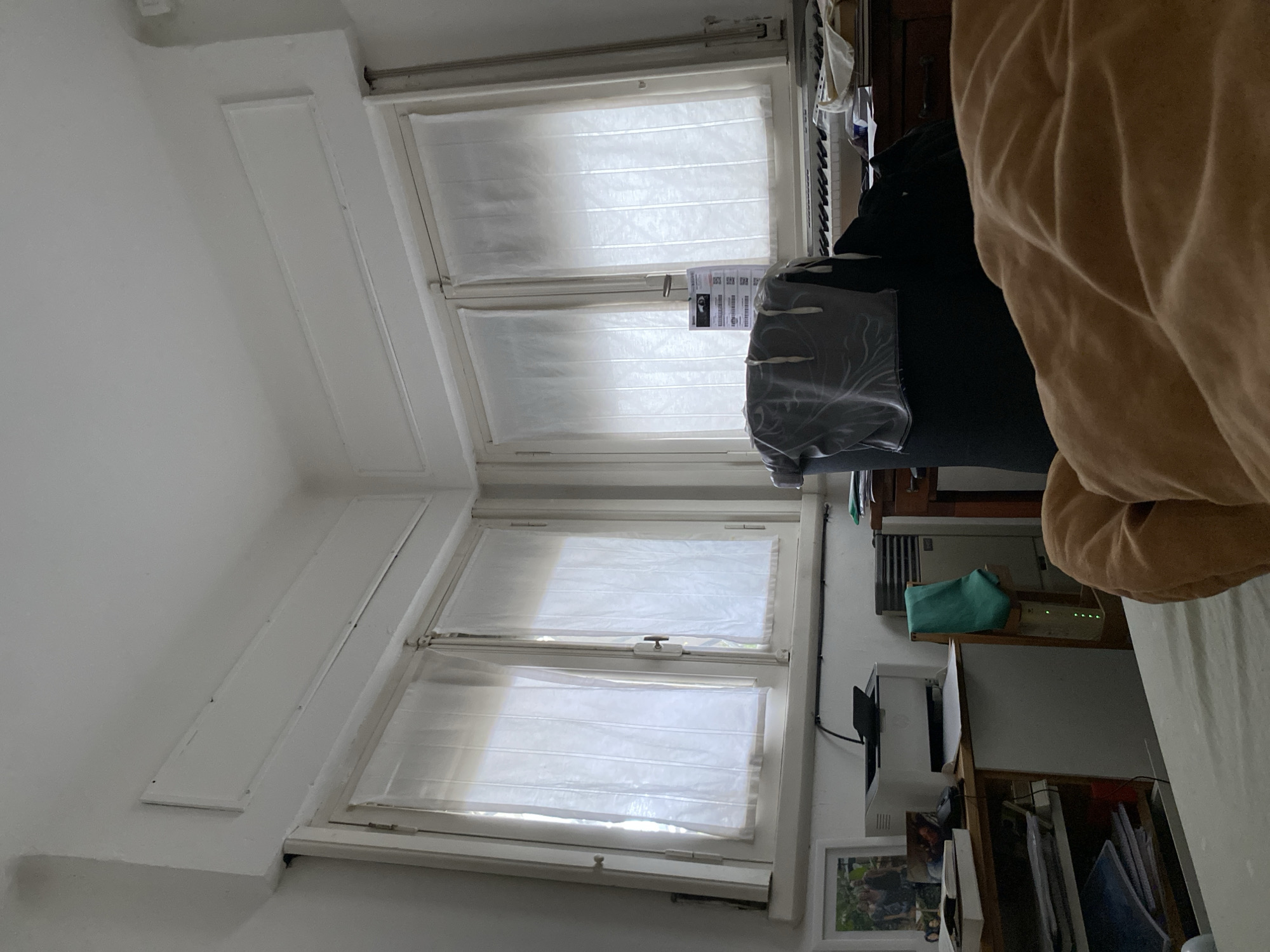 The image depicts an indoor scene, likely a bedroom or study, with a prominent corner of the room featuring two adjacent windows. The windows are covered with translucent white curtains, allowing diffused light to enter. Above the windows, a white decorative molding runs along the ceiling.
In the foreground, a soft, brown blanket is partially visible, suggesting someone is lying down or has recently been there. To the left, a shelf unit holds various items including books, a framed photograph of people, and a white printer. To the right of the printer, a grey vertical heater unit with a green towel hanging on it is positioned.
Further to the right, a wooden desk is visible with a keyboard of a musical instrument on it. A black office chair with a grey garment draped over its back is situated in front of the desk. Papers and other office supplies are scattered on the desk.
The overall impression is of a lived-in, functional space. The location in Martínez, Argentina, is not visually confirmed by any specific landmarks or identifiable elements within the frame.
