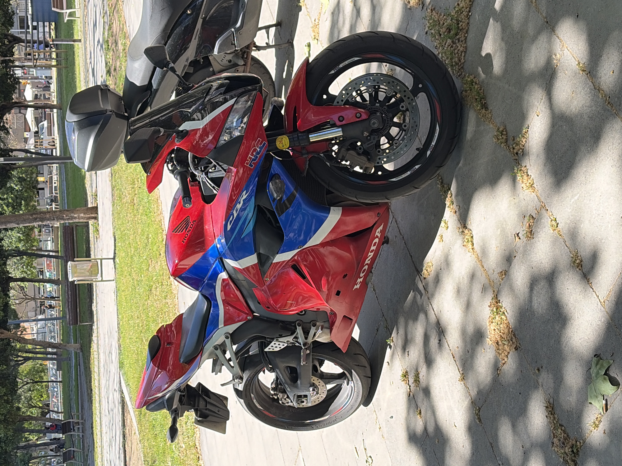 A Honda CBR motorcycle with a red, white, and blue color scheme is parked on a paved surface. The background shows trees, grass, benches, and buildings suggesting a park or public area. The location appears to be in a sunny outdoor setting. Based on visible details and landmarks, the location is within the city of L'Hospitalet de Llobregat, Spain.
