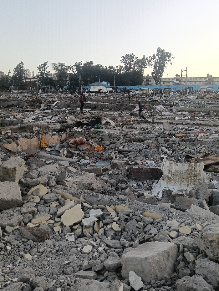 A vast expanse of rubble dominates the scene, filled with fractured concrete, stone fragments, and debris. The sky is overcast. Distant trees and buildings line the horizon. Several figures traverse the ruins, likely navigating through the devastation. The foreground showcases larger chunks of broken material, while the background depicts a flattened landscape of smaller pieces. The scene evokes a sense of destruction and urban decay.