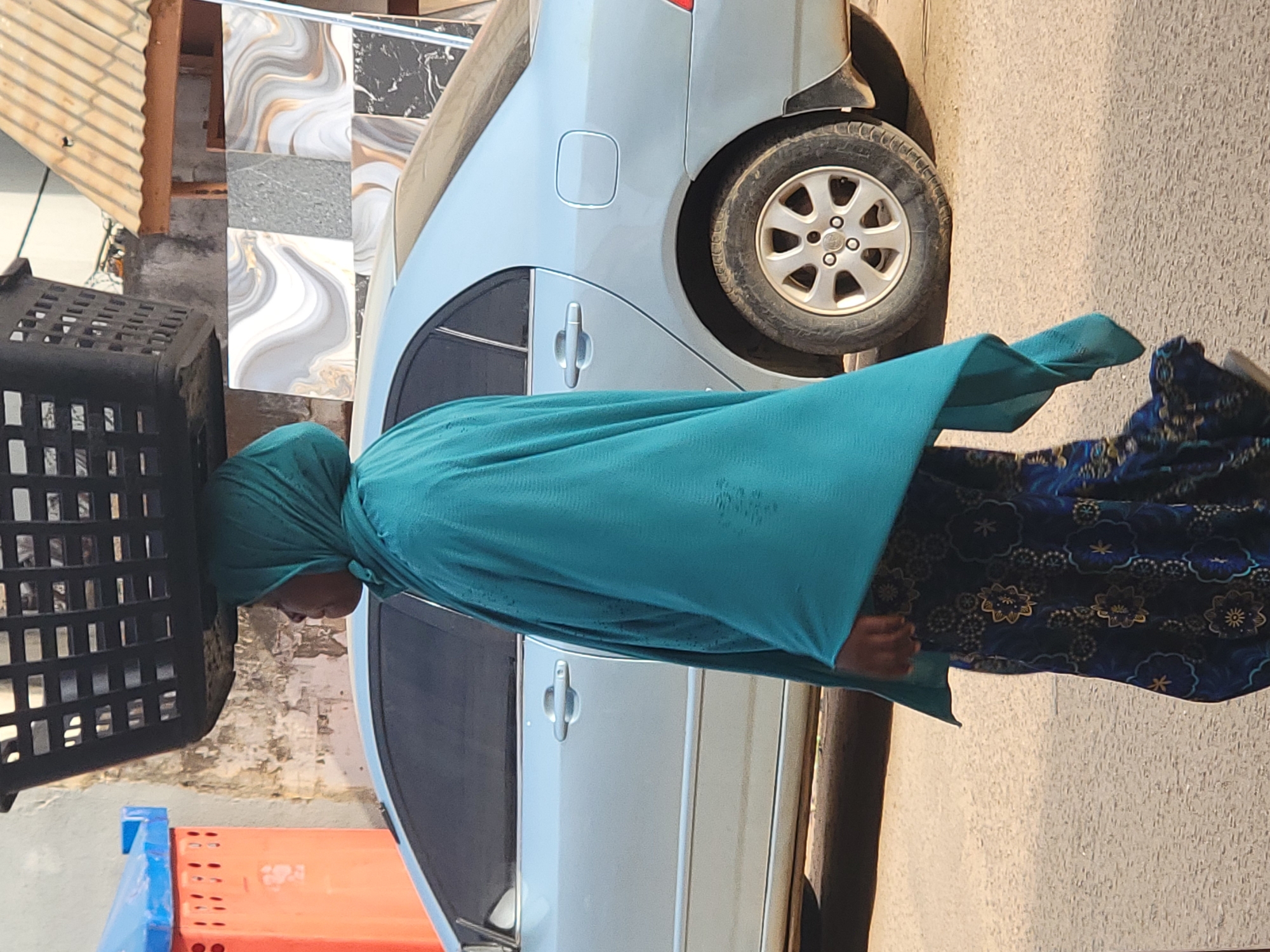 A woman in Maiburiji, Nigeria, with a teal head covering and blue floral skirt walks along a street, carrying a black plastic basket on her head. To her right is a parked silver car, and behind the car are stacks of construction materials. The background includes a building with a corrugated roof.
