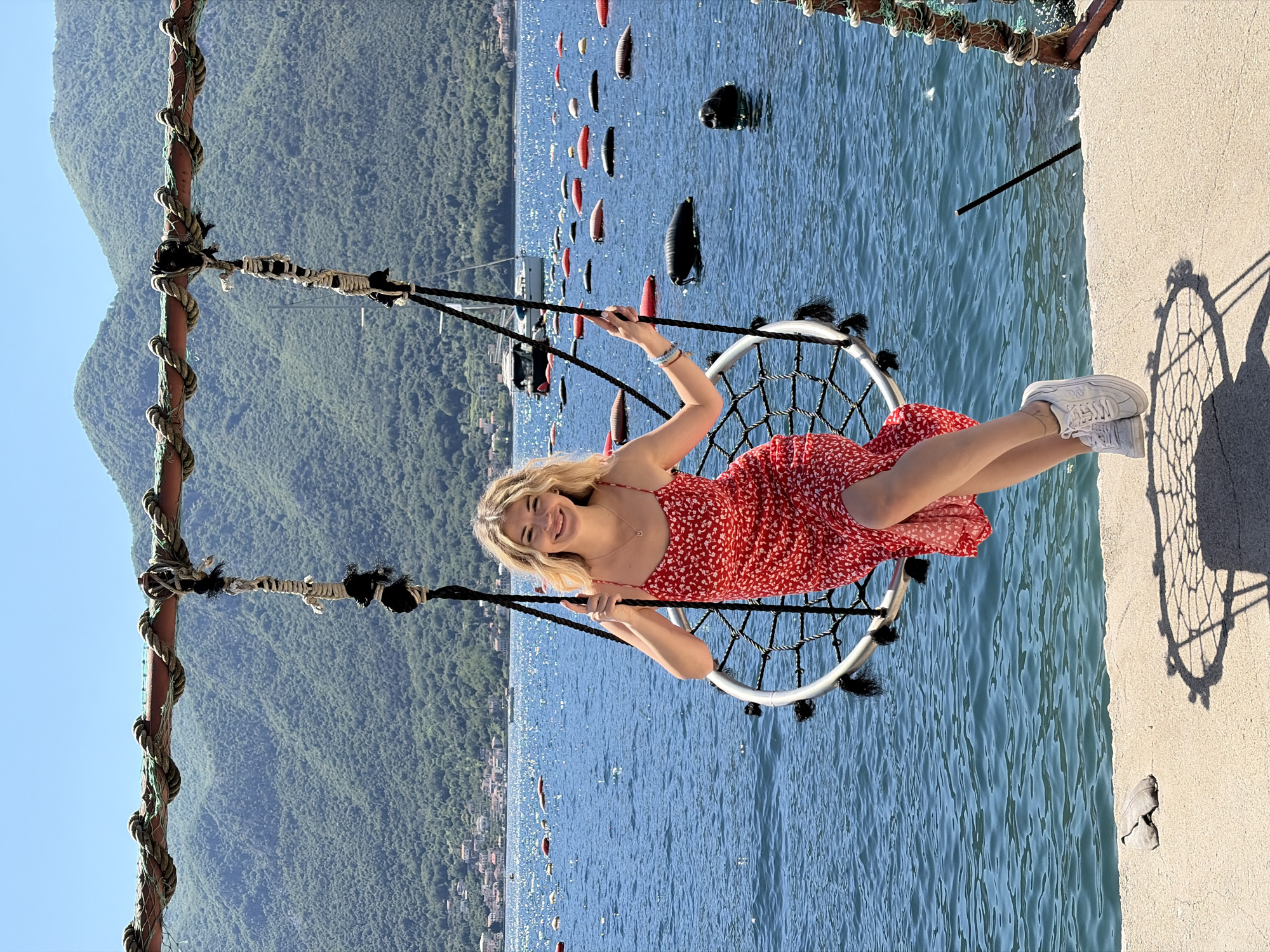 A woman in a red floral dress and white sneakers is seated in a rope swing suspended over blue water. She is holding onto the ropes of the swing and smiling at the camera. The swing is suspended from a rusted metal frame with thick ropes wound around it. In the background is a body of water with several buoys floating on the surface, as well as a mountain covered in green trees. The location is Dražin Vrt, Montenegro. The scene is well-lit, suggesting it is daytime. There is a concrete area on the right side of the frame where the swing is casting a shadow.
