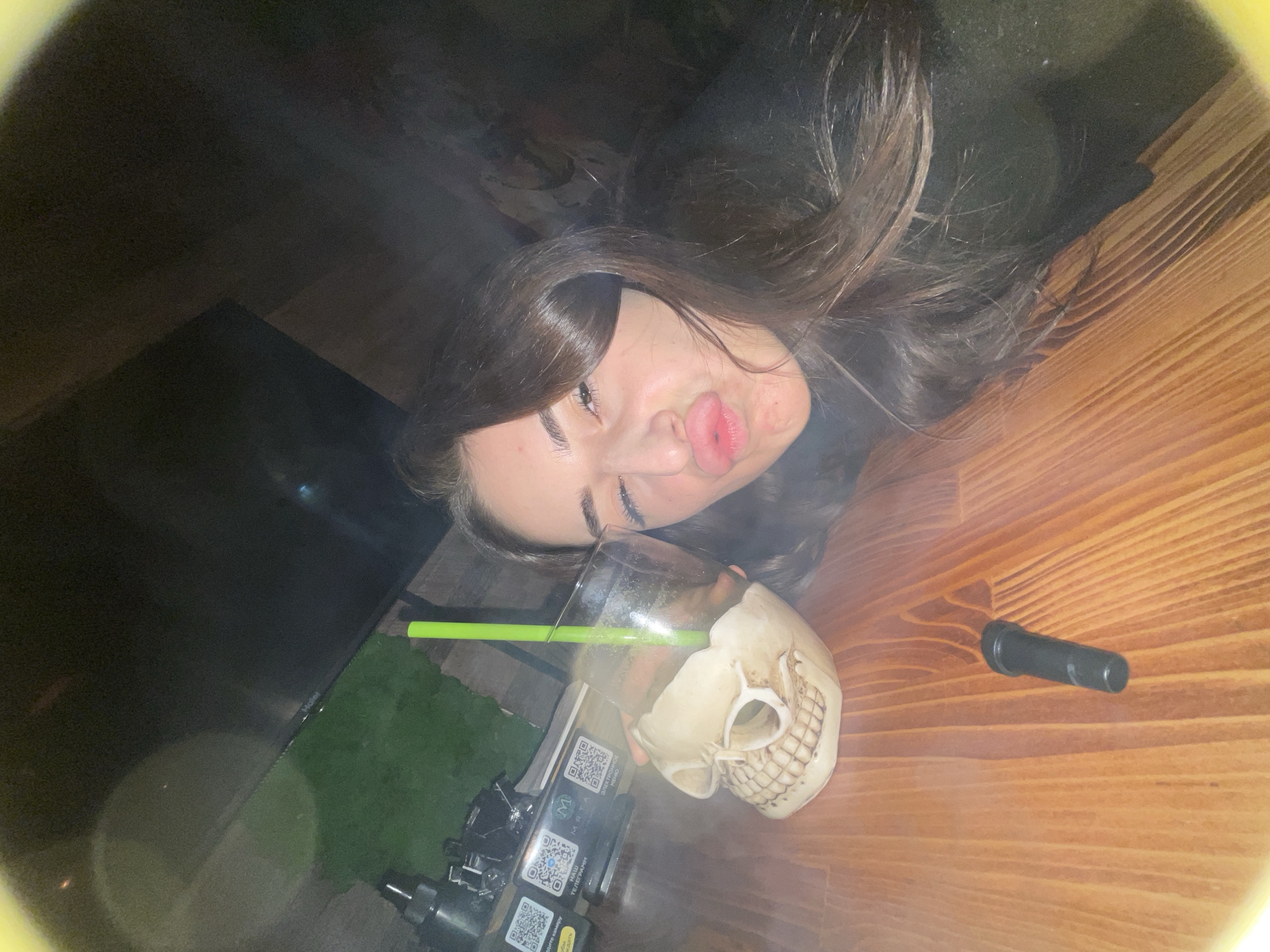 A young woman is oriented toward the viewer with a "kissing" facial expression. She is holding a skull-shaped glass with a green straw, likely containing a beverage. Part of a hookah and related accessories are visible. The location is Moscow, Russia. The woman is positioned indoors, possibly in a private residence, given the background of wooden flooring, a television, and other furniture. The lighting appears to be dim. An additional black object (possibly another part of the hookah) is also visible.