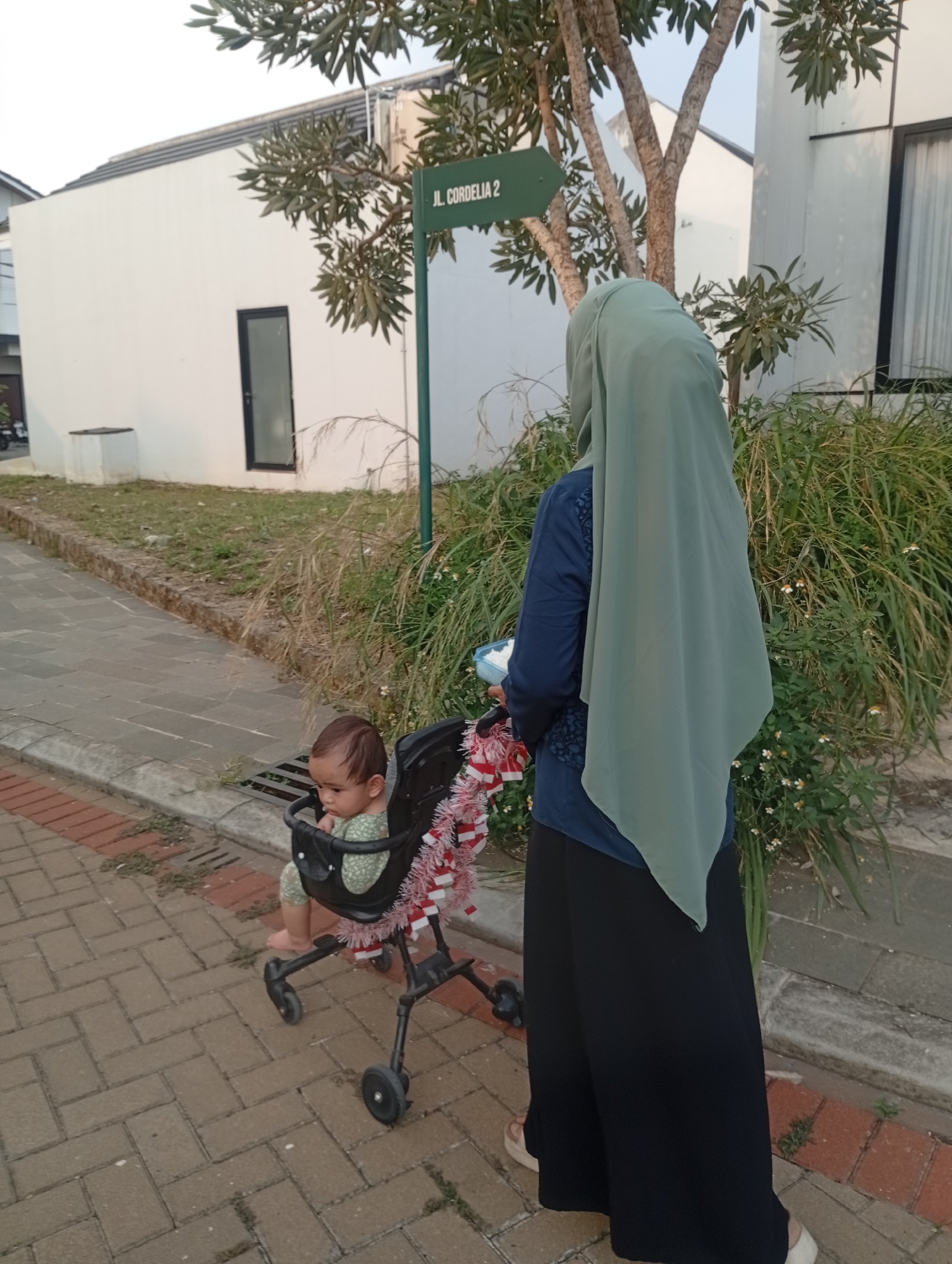 In Kemang, Indonesia, a woman wearing a light green hijab and a dark blue top with a patterned overlay stands in front of lush greenery and a white building. She holds a light blue object in her right hand. To her left, a baby sits in a black stroller adorned with red and white garland. The stroller is positioned on a brick pathway, adjacent to a grassy area. A green street sign indicating "JL. CORDELIA 2" is visible behind the woman and the stroller, mounted on a pole. The environment suggests a residential or suburban setting.