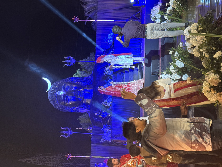 The image depicts an elaborate evening event, likely a wedding or a pre-wedding ceremony, characterized by dramatic lighting and a prominent Hindu theme. The scene is set outdoors at night, with the primary illumination coming from artificial blue and white spotlights.

The central and most striking feature is a large, dark-colored statue of the Hindu deity Shiva, positioned towards the left side of the frame. The statue is intricately detailed, featuring a crescent moon on its head, an urna (third eye mark) on its forehead, and long, flowing hair. It is bathed in a vivid blue light, giving it an ethereal glow. Nearby, decorative tridents (Trishuls), a symbol associated with Shiva, are visible as part of the overall setup.

In front of the Shiva statue, a couple, presumably the bride and groom, are positioned. The groom, standing closer to the statue, is dressed in a light-colored, ornate sherwani, possibly white or silver, and wears a matching turban or headpiece. The bride, slightly to the right of the groom, is seen from behind, wearing a traditional red and gold lehenga or saree, adorned with intricate embroidery.

Several other individuals are present at the event. A man in a dark purple shirt and light-colored pants stands just behind the couple, actively taking a photo or video with a camera or phone, facing towards the couple. In the foreground, another man, seen from behind, wears a distinctive patterned jacket (possibly abstract or camouflage-like) over a dark shirt. His arm is raised, suggesting he is also interacting with the scene, perhaps taking a photo or gesturing. Partial views of other guests in traditional attire are visible in the foreground and background, indicating a gathering.

The setting includes decorative elements such as lush white floral arrangements, visible on the right side of the image, possibly adorning pillars or a stage. The background is mostly dark, with visible beams of light cutting through the air, highlighting the smoky or misty atmosphere often created by stage effects. A structural framework, possibly part of a tent or stage, is faintly visible on the far left. The overall ambiance is grand and celebratory, steeped in cultural tradition. No text is visible in the image.