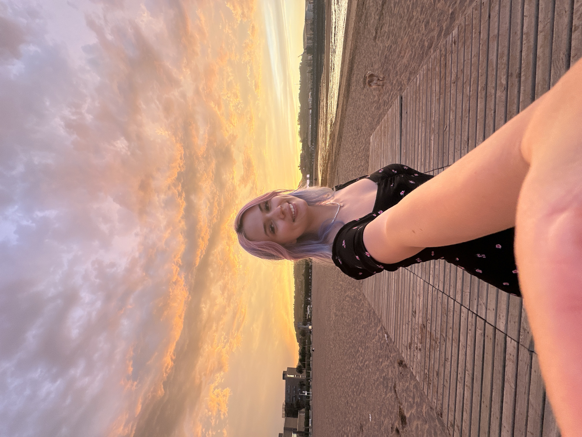 In Gijón, Spain, at dusk, a young woman with pink and purple dyed hair takes a selfie on a wooden boardwalk that leads onto a sandy beach. She is wearing a black dress with a floral pattern and a pearl necklace. The sun is setting, casting an orange and purple glow across the sky, illuminating the scattered clouds. In the background, buildings and a distant hill are visible. A small dog is walking on the beach to the right of the boardwalk. The tide is low, exposing wet sand near the water.