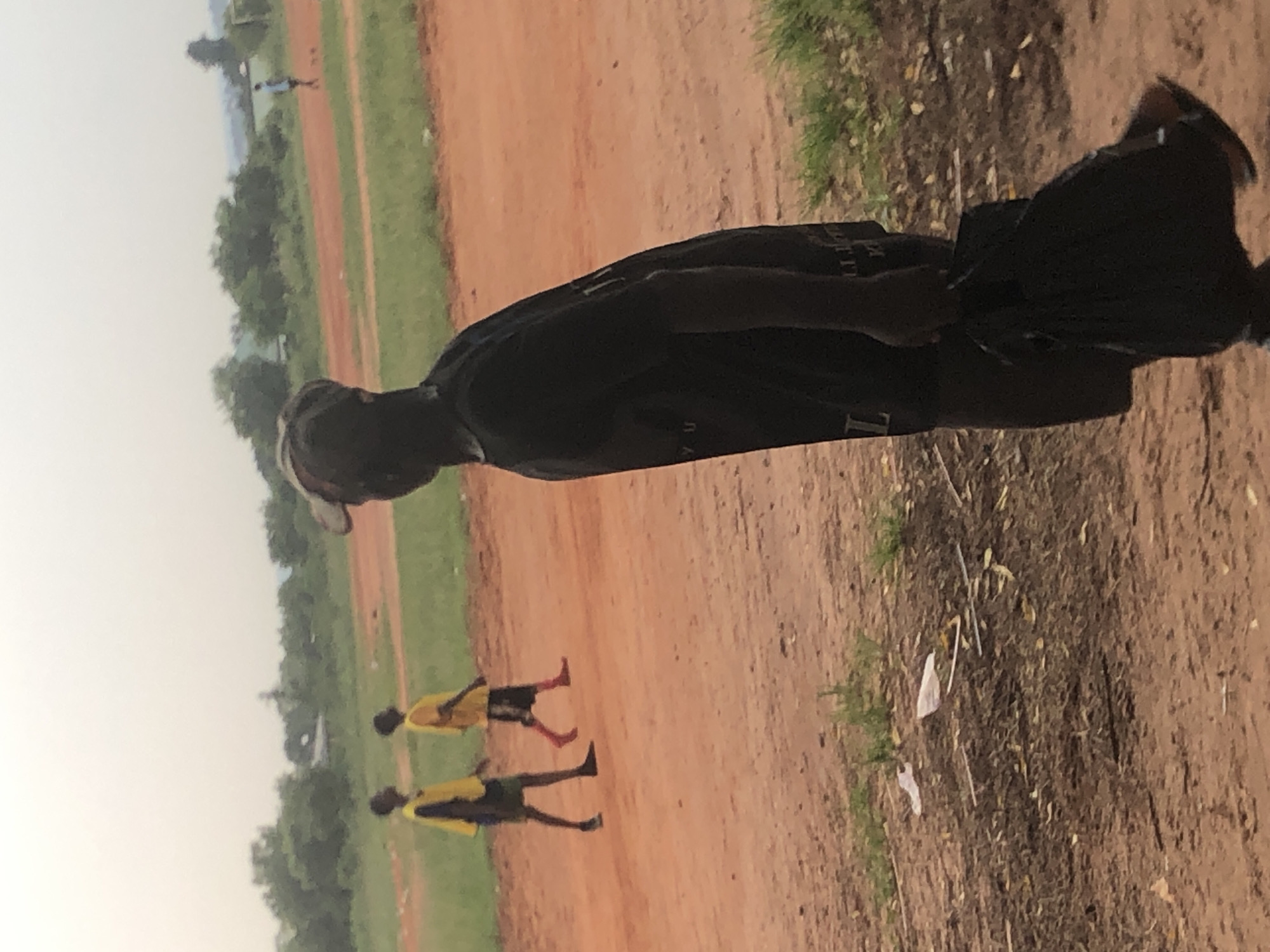 On an overcast day in Ukwu-Abwa, Nigeria, two young people wearing yellow shirts and dark shorts are running down a dirt path. An additional individual is visible in the distance. A person in a black garment is in the foreground. The surrounding environment includes dirt, grass, and scattered debris. Vegetation and buildings are situated in the background.