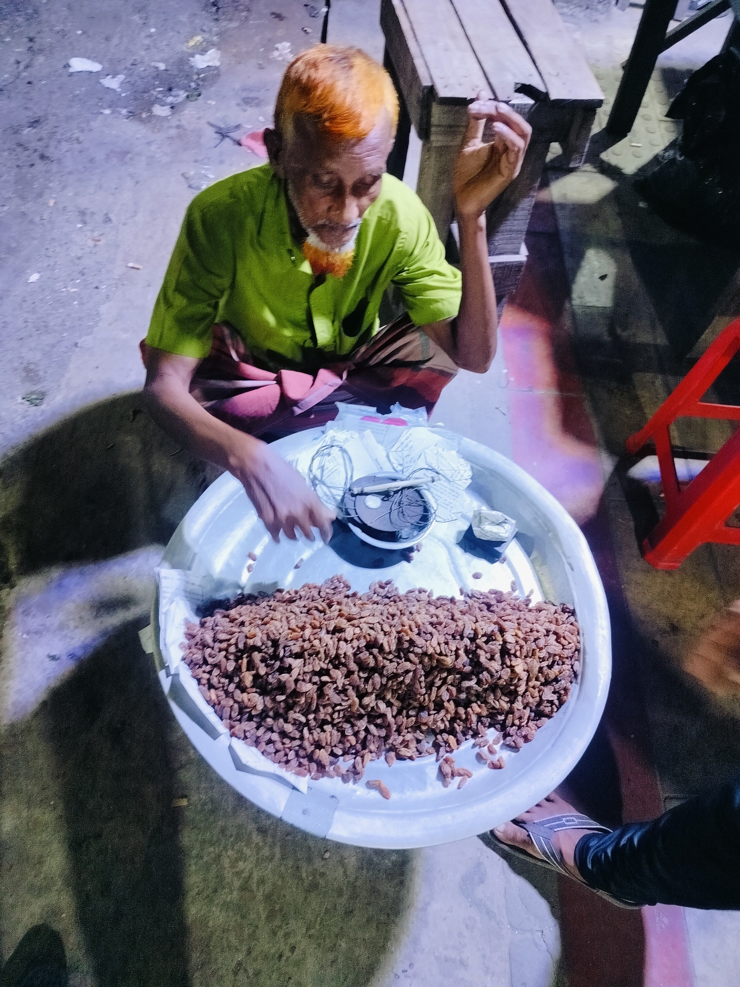 In Dhaka, Bangladesh, an elderly man with dyed orange hair and beard is sitting on the ground next to a large, shallow, white metal tray overflowing with raisins. He wears a lime green shirt and a reddish-brown lungi. His right hand is raised near his face, and his left hand is resting on the edge of the tray. The tray contains not only the raisins but also some crumpled paper and a small dark container. The ground appears to be concrete, with some scattered debris. In the background, a wooden bench and a red plastic chair are visible, illuminated by artificial light that casts long shadows. A portion of a person's leg, wearing black pants and a sandal, is visible in the lower right corner of the frame, suggesting the perspective of someone standing nearby.