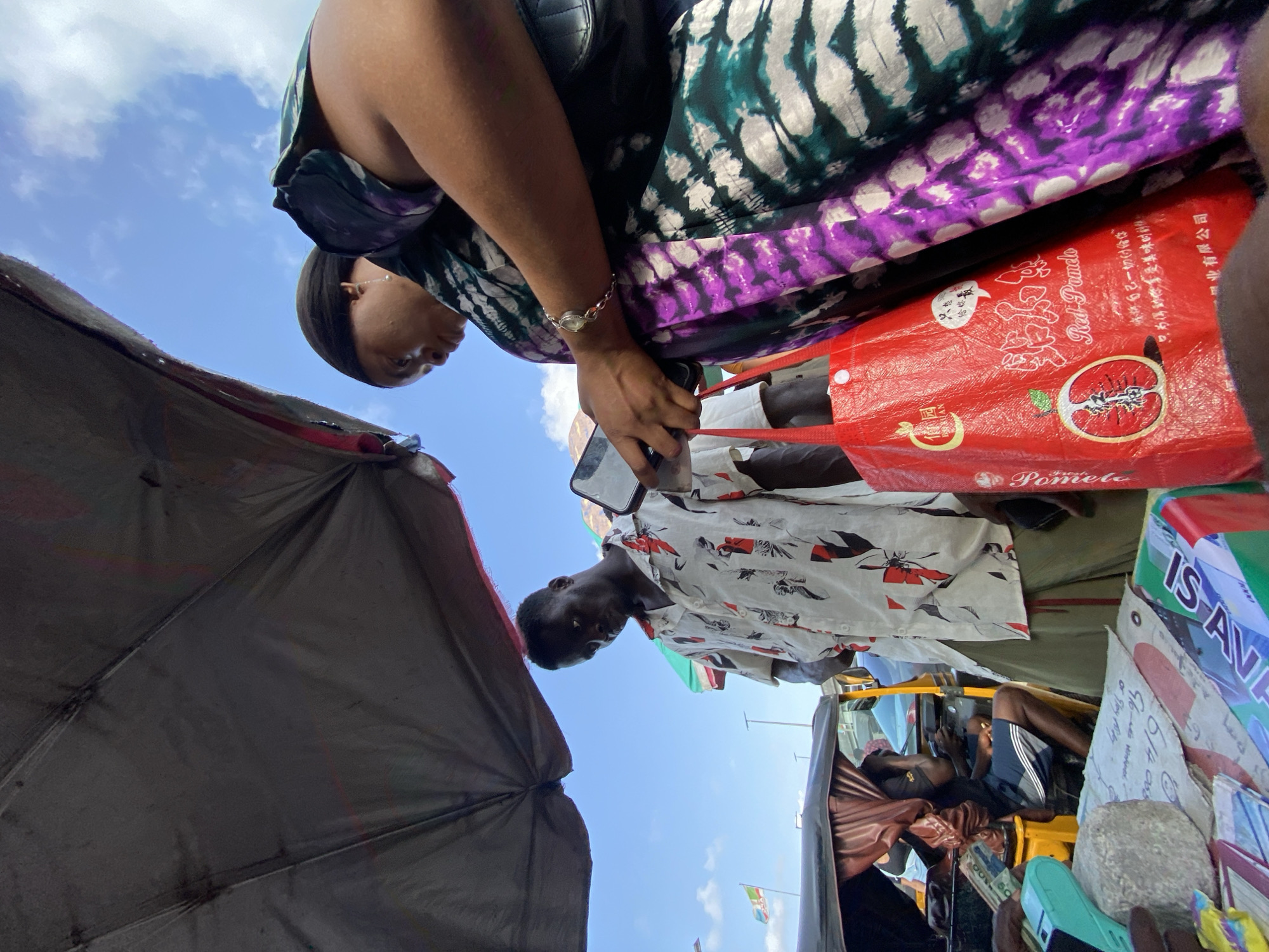The image, taken from a low angle, depicts a street scene in Lagos, Nigeria. The sky is blue with scattered clouds. In the foreground, a gray umbrella offers shade. Two people are visible: a woman wearing a dress with green, black, and purple patterns, holding a red shopping bag with Chinese writing and the image of a pomelo on it, and a man in a white shirt with red and black designs. The woman is looking down, presumably at something in the bag or her phone. The man appears to be looking toward the person taking the picture. A yellow "Keke Napep" tricycle taxi is visible in the background, with a driver in the vehicle. Advertisements and signage are visible, including a sign that says "IS AVA." The overall scene suggests a busy marketplace or commercial area.