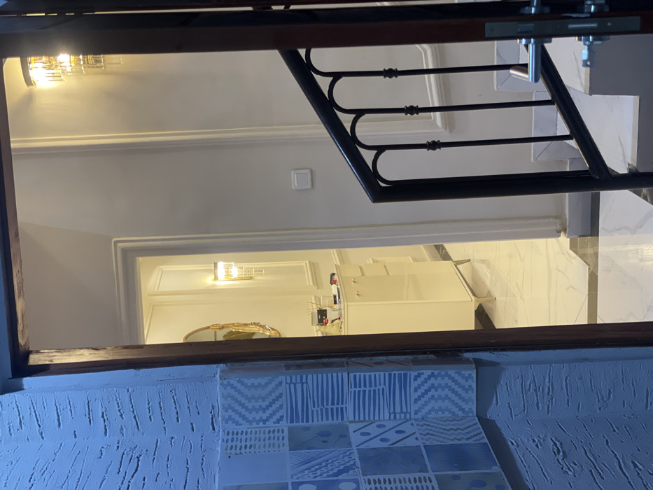 The image, rotated to its correct upright orientation, depicts an interior view within a building, possibly a residence or commercial space. In the immediate foreground, a dark wooden beam or railing horizontally frames the scene. Below this beam, a wall features a textured, light blue-grey plaster finish, partially covered by decorative tiles arranged in a grid pattern. These tiles display various geometric designs, including chevrons, stripes, and abstract shapes, in shades of blue, white, and grey.
Beyond the foreground railing, a black wrought iron balustrade with ornate scrollwork extends upwards, suggesting a staircase or an upper-level landing. The surrounding walls are painted a light color, likely off-white, and feature classic decorative moldings. A white electrical switch is mounted on one of these walls.
Further back in the scene, a modern wall sconce with a gold finish casts light on an upper wall. Below it, a white doorway with trim leads into an adjacent room. Inside this room, another similar wall sconce is visible, alongside a golden-framed oval mirror mounted on the wall. A white rectangular unit, possibly a refrigerator, cabinet, or an electrical power box with visible wiring, stands within this room. The floor of the lower level, visible towards the right, is covered with glossy, marble-patterned tiles, leading to what appears to be the bottom of a staircase.
No people are present in the image, and no specific activity is occurring. The lighting suggests an indoor setting illuminated by artificial sources, possibly indicating evening or a reliance on interior lighting. No discernible text is visible anywhere in the image. The architectural elements appear modern and well-maintained.