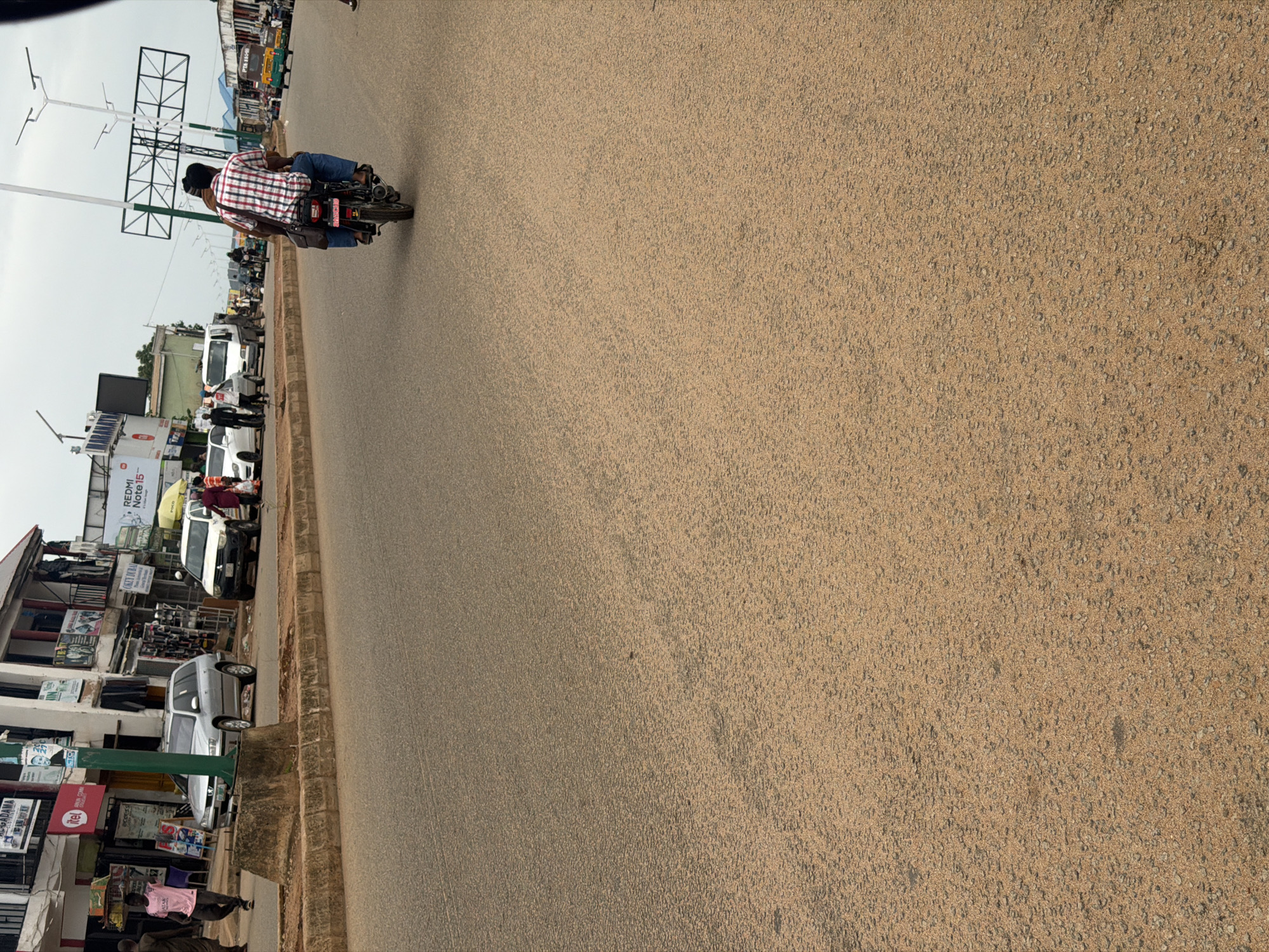 The image depicts a street scene in Bukuru, Nigeria. The perspective is from a slight elevation, looking down onto a road with vehicles and buildings lining one side. The dominant activity is traffic, with several vehicles present including cars and a motorcycle. On the motorcycle, two people are riding, the passenger wearing a red and white checkered shirt.
The scene appears to be during the daytime, with overcast or hazy skies suggested by the diffused light. The road surface is paved with asphalt and has a rough texture. Buildings on the left side of the road show commercial activity, with shops and signs visible. One sign prominently displays "REDMI Note 15". Other signs are partially obscured or in a language that is not immediately identifiable.
The overall atmosphere is that of a typical urban street with ongoing commerce and transportation. The image captures a moment in the daily life of the location, focusing on the movement of people and vehicles within the built environment.
