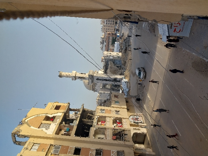 The image displays a wide-angle view of a street scene in Khan Yunis, Palestinian Territory, characterized by extensive urban destruction under a clear sky. On the left, a multi-story residential building exhibits severe structural damage, with large sections of its facade missing, exposing internal rooms, broken windows, and some visible belongings like laundry on a balcony. In the midground, a mosque with a domed roof and a tall, slender minaret stands prominently, showing visible damage to its structure and surrounded by debris. The paved street below, marked with white lines, is occupied by approximately 15-20 individuals, including adults and children, walking in various directions. A white van is present on the street. The scene is framed on the right by a partial view of another damaged building, with multiple overhead utility wires traversing the upper portion of the frame. Further into the background, additional damaged buildings and piles of rubble are visible, indicating widespread destruction.