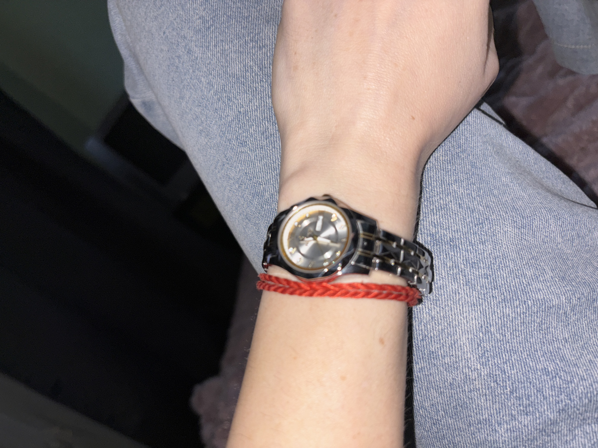 A close-up shot focuses on a person's arm and wrist, adorned with a red braided bracelet and a silver-toned watch. The arm is resting on light blue denim material, likely pants or a jacket. The background is dark and out of focus, with hints of what appears to be a brick surface in the lower right corner. The lighting suggests an indoor setting, possibly at night. No specific location in the city of Unknown, Russia, can be determined from the visual evidence.