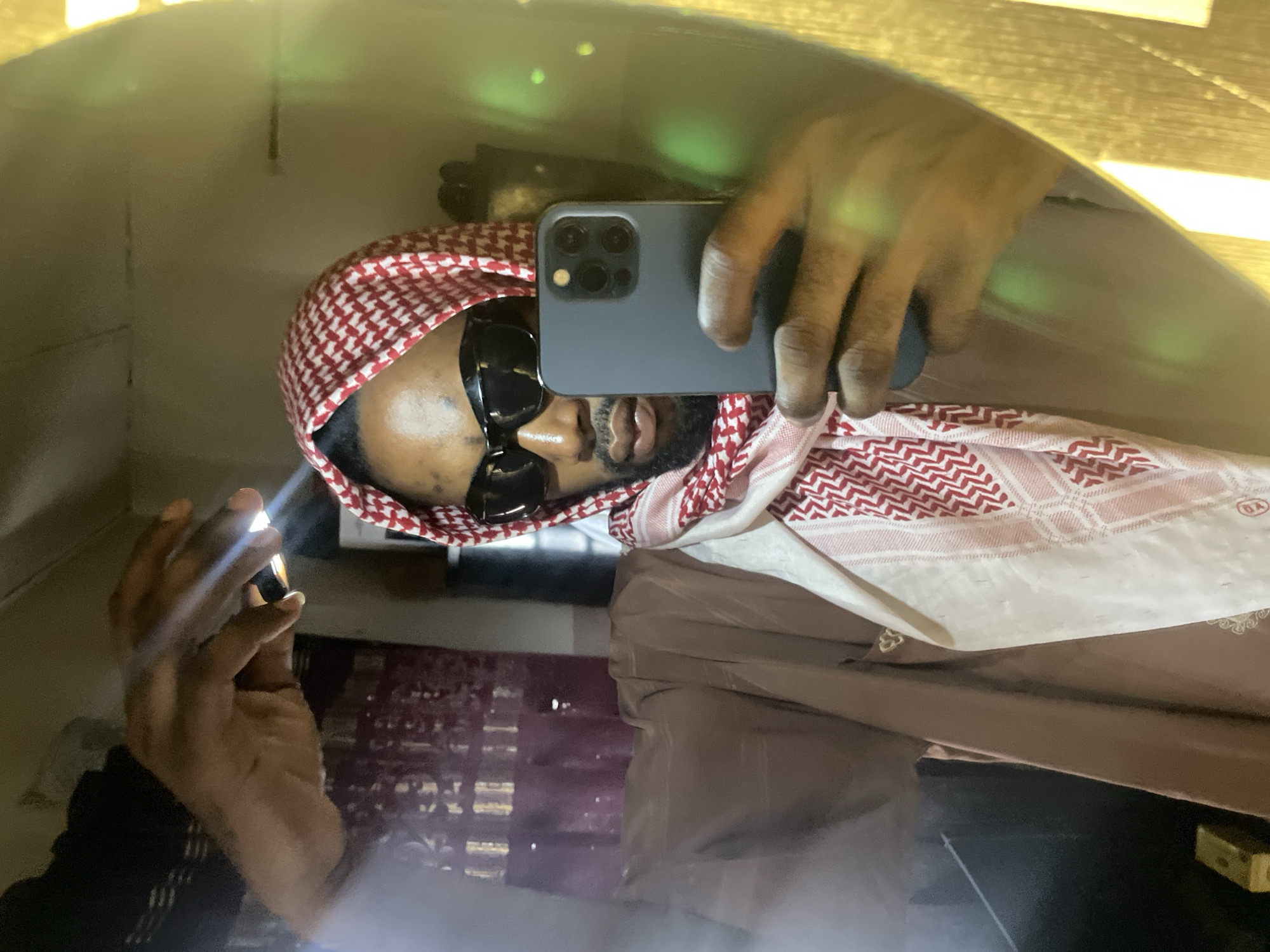 A selfie reflection shows a person wearing a red and white patterned headdress and dark sunglasses, holding a gray smartphone. The reflection suggests the person is inside an enclosed space, possibly a shop or a room, with a dark or mirrored surface creating the reflection. No specific details pinpoint the location as Lokoja, Nigeria. The image lacks identifiable landmarks or cultural elements that would confirm this location.
