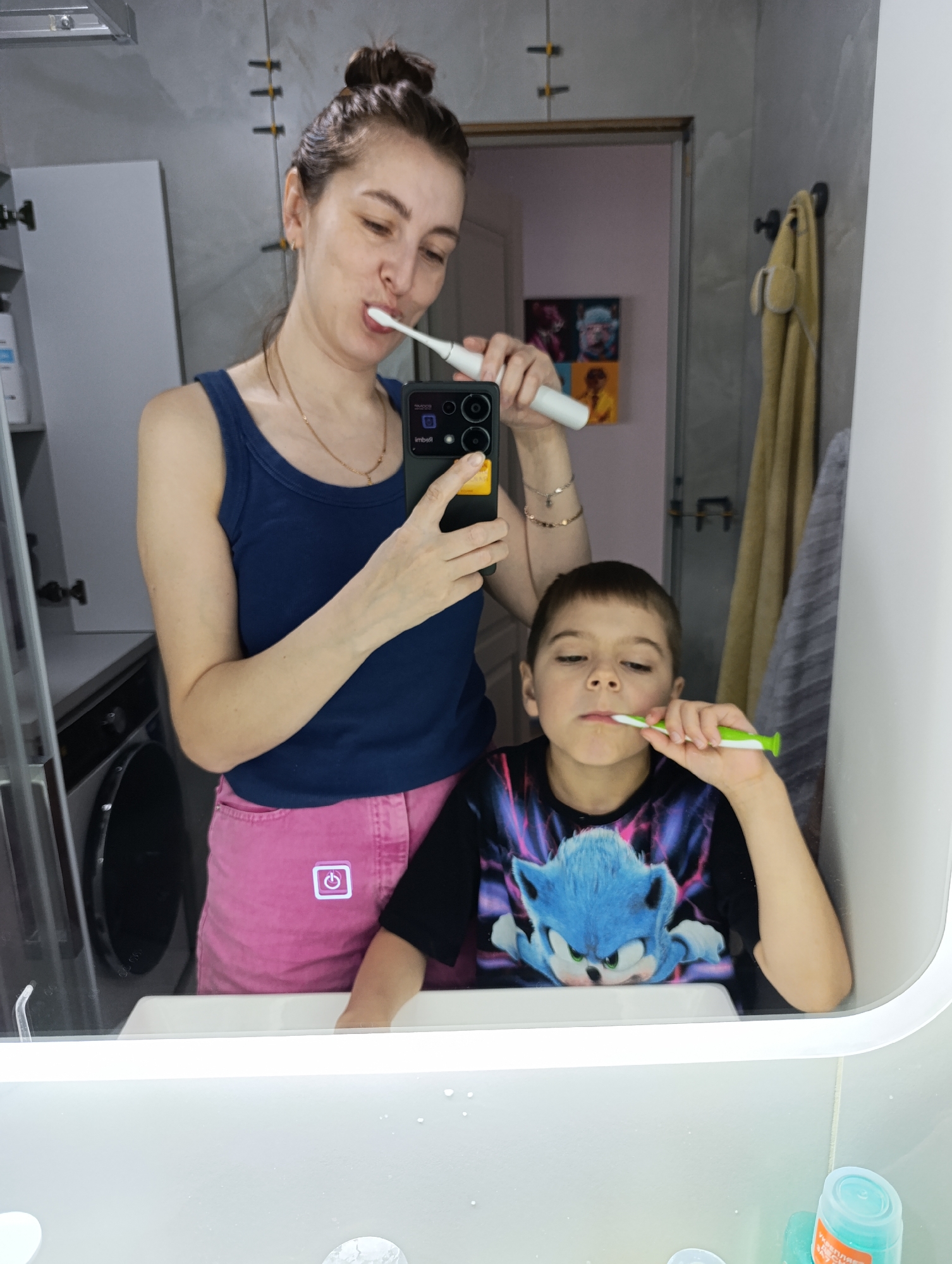 An adult female and a young male child are observed in a bathroom in Chaikovsky, Russia, reflected in a large, illuminated mirror. The female, with brown hair styled in a bun, is wearing a navy blue tank top and pink shorts. She is actively brushing her teeth with a white electric toothbrush while simultaneously holding a black smartphone in her left hand, utilizing its reflection to capture the image. She wears a gold chain necklace and a bracelet on her right wrist. To her right, a male child, estimated to be between 6-10 years old, with short brown hair, wears a black t-shirt featuring a blue Sonic the Hedgehog graphic. He is also brushing his teeth with a green and white manual toothbrush, focused on the activity. Both subjects are positioned in front of a white sink, with water droplets visible on the lower edge of the mirror. The background reflection reveals a grey front-loading washing machine to the far left, white wall cabinets, and a set of three framed artworks depicting stylized, brightly colored animal faces. Two towels, one beige and one grey, hang on hooks to the right side of the mirror. A teal bottle and a clear cup are present on the sink counter in the bottom right corner of the reflection. The bathroom features light grey, possibly tiled, walls.