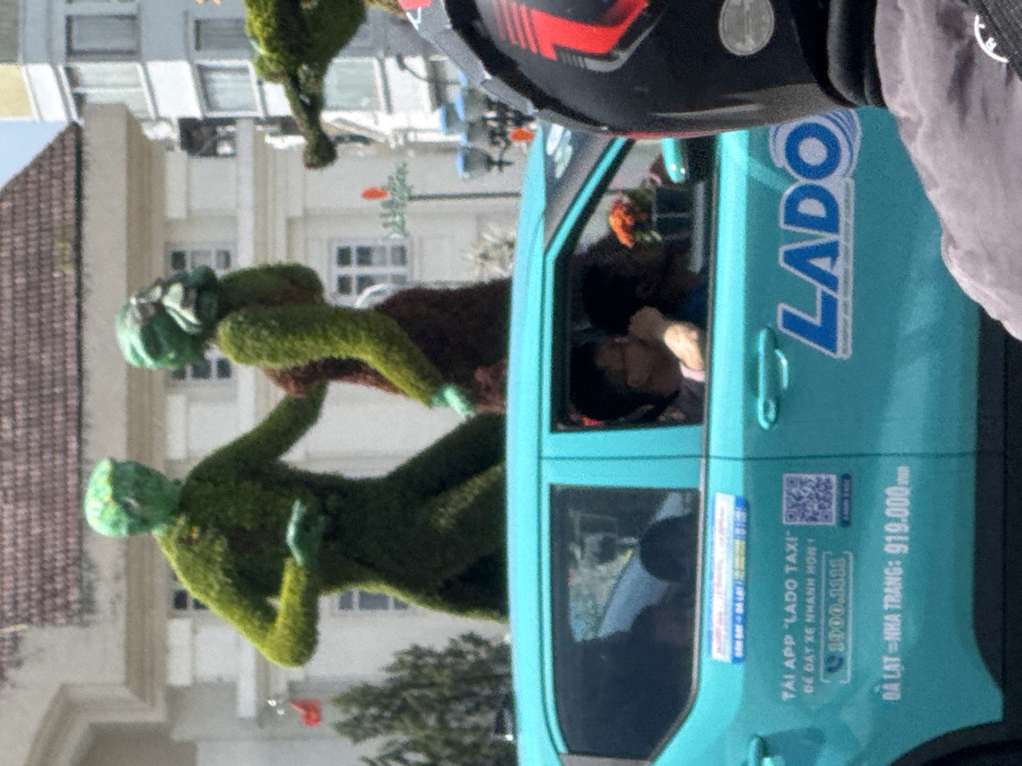 In Da Lat, Vietnam, a turquoise taxi with "LADO" branding and a QR code is parked. Above the taxi, two large, green, topiary figures, resembling aliens or mythical creatures, are positioned against a building with a tiled roof. The figures are covered in moss or greenery, with one figure in the foreground and another slightly behind it. The one in the foreground has a rounded head and appears to be reaching out with its right arm. The second figure, to the right, is partially obscured. Inside the taxi, passengers are visible through the windows, including a person in the front passenger seat and another in the back. A person wearing a black motorcycle helmet with red accents is partially visible in the foreground on the right. The background includes trees and other buildings.