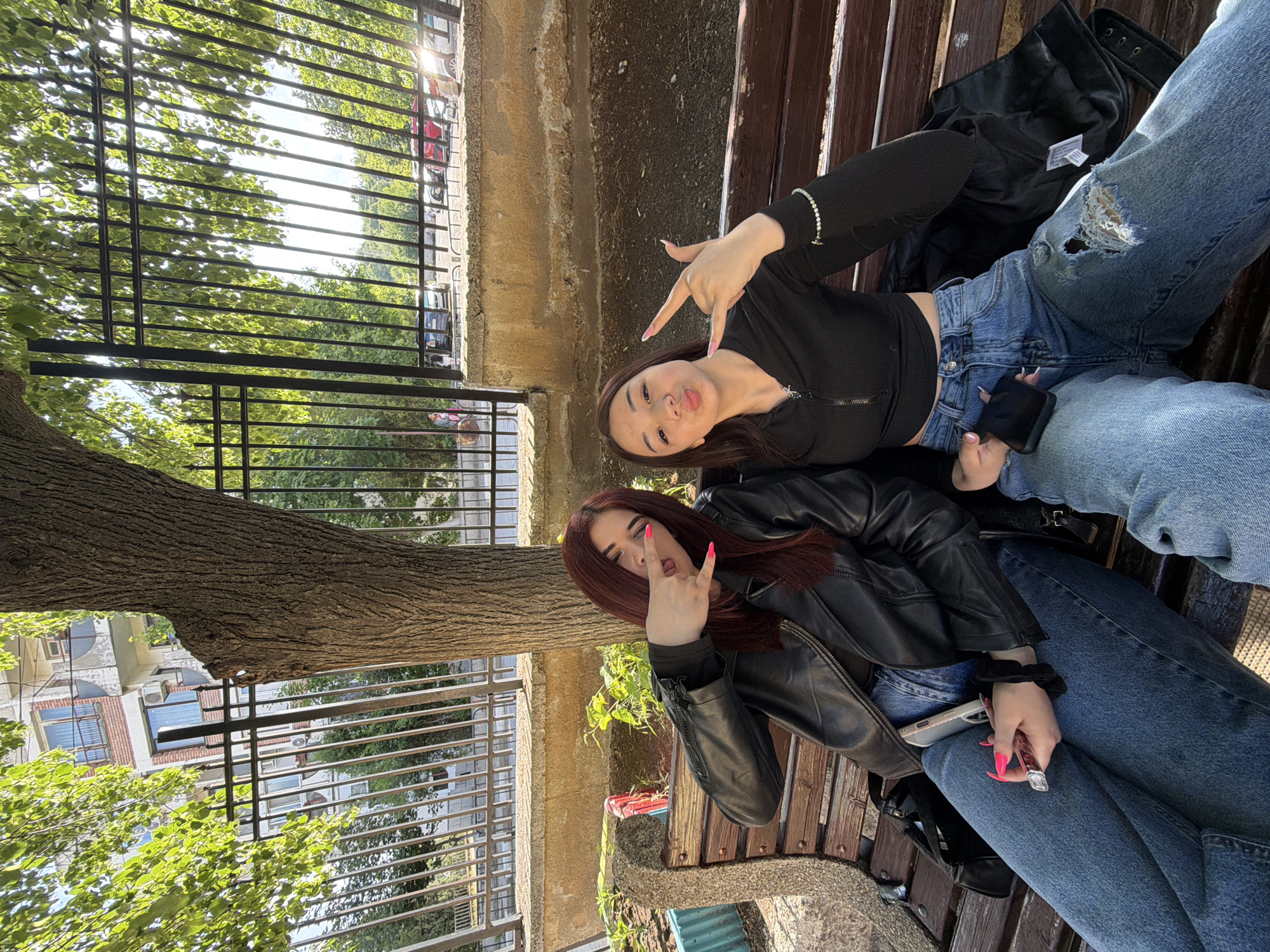 This outdoor image captures two young women posing playfully on a wooden park bench in Bobov Dol, Bulgaria, under bright daylight. The camera appears to be tilted, capturing the scene from an elevated angle.
The woman on the left, with dark brown hair, wears a black long-sleeved, zip-up top and light blue ripped jeans. She flashes a "rock on" hand gesture with her right hand, displaying long, pink manicured nails, and has her lips slightly puckered towards the camera. A silver bracelet is visible on her left wrist.
Beside her, on the right, sits another woman with reddish-brown hair, clad in a black leather jacket over a dark top and blue jeans. She also makes a "rock on" gesture with her right hand, her bright pink nails contrasting with her jacket, and her tongue playfully extended. She holds a smartphone and a small, clear lighter in her left hand.
The bench they are seated on is made of dark wooden slats supported by concrete. Behind them, a large tree trunk is visible on the left, with vibrant green leaves filling the upper part of the frame. A black metal fence runs horizontally through the mid-ground, separating the park from the background. Beyond the fence and foliage, several multi-story buildings, some with air conditioning units, and parked cars can be seen, indicating an urban setting. A black jacket rests on the bench between the two women, displaying a white, unreadable tag. The scene conveys a casual and friendly atmosphere.