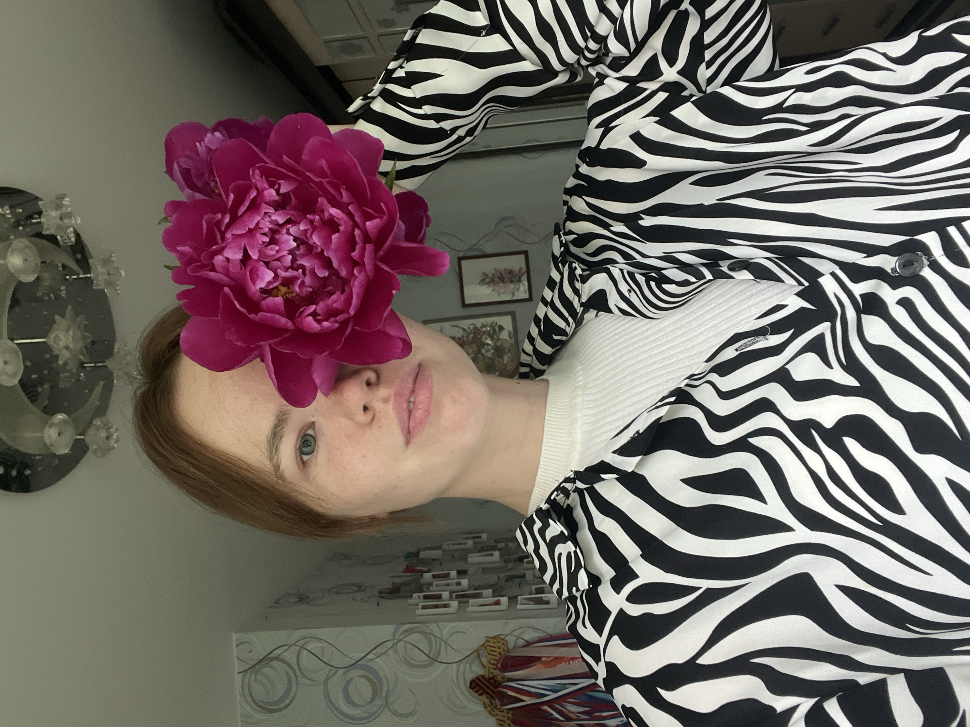 The image shows a young woman with light skin and reddish-blonde hair holding a large, vibrant pink peony flower up to her face, obscuring her right eye and part of her nose. She is wearing a white ribbed top underneath an open black and white zebra-print shirt. Her expression is neutral. In the background, there is a patterned wallpaper with a faint floral design, a chandelier with multiple crystal-like elements on the ceiling, and framed pictures on the wall. To the left, a rack with medals or awards is visible. The environment appears to be indoors, likely a residential room. This scene is not specifically identifiable as being in Chaikovsky, Russia, as there are no distinctive landmarks or cultural elements present that would place it in that specific city.