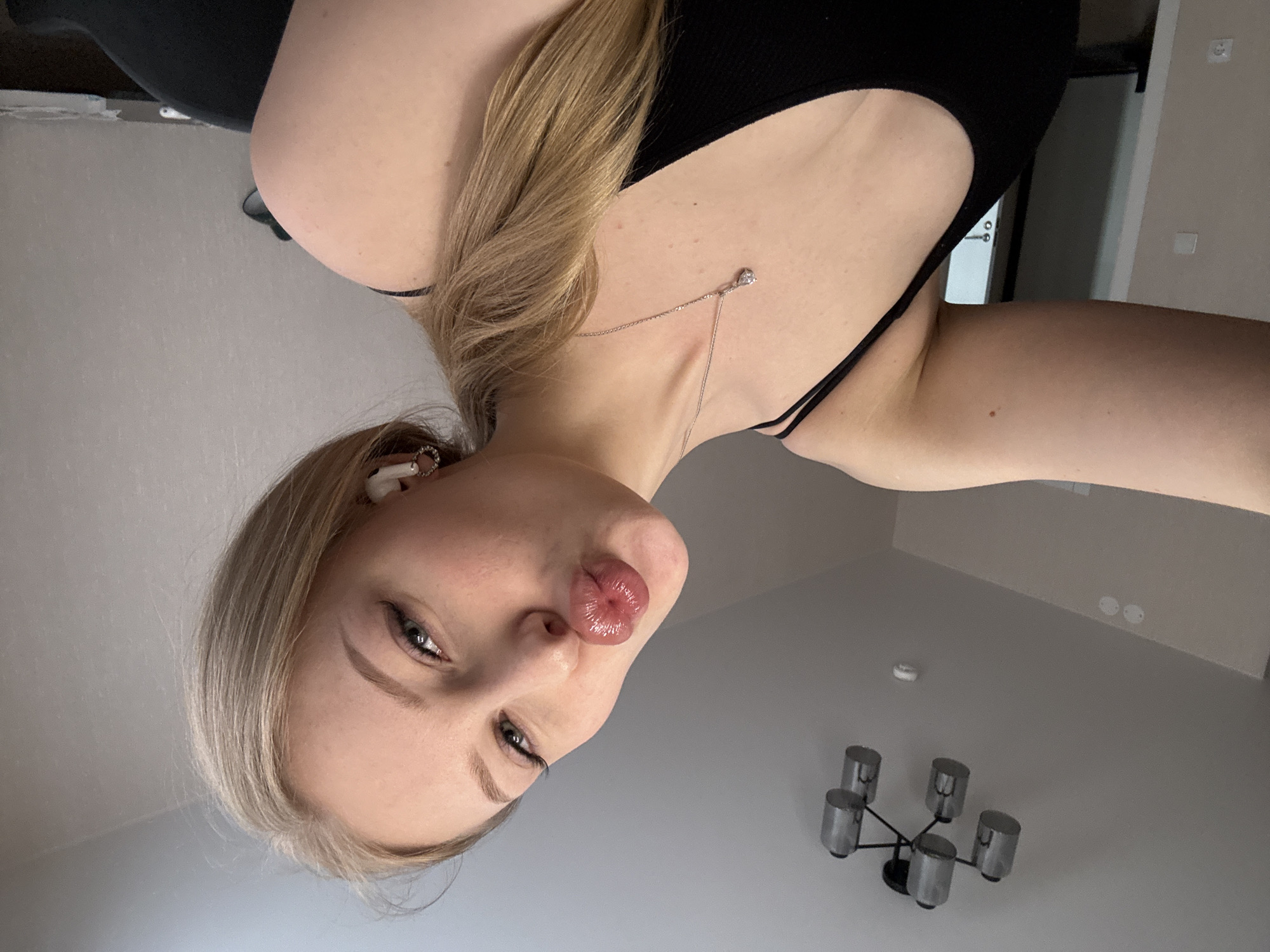 A low-angle selfie of a young woman with blonde hair, wearing a black top and a silver necklace, is presented in portrait orientation. She has an earbud in her left ear and is puckering her lips. The background includes a gray wall, a chandelier with five light fixtures, and a door. The photograph was taken indoors, location is Perm, Russia.