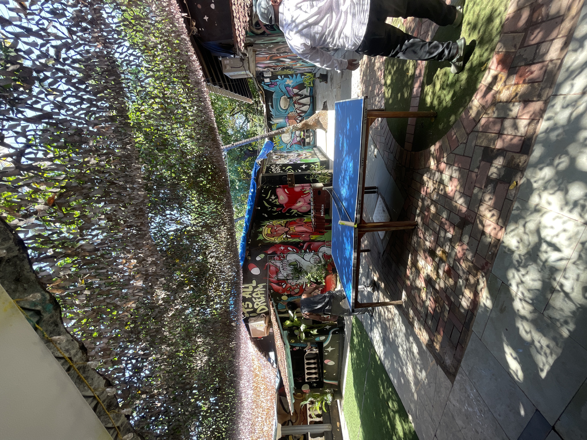 The image depicts an outdoor recreational area adorned with vibrant graffiti art. In the foreground, a blue ping pong table is set up on a paved area. To the right of the table, a person wearing a white shirt and dark pants is walking away from the camera. To the left of the table, another person in a gray tank top and pants stands near the table. The background features a building covered in colorful graffiti, with visible plants and trees. Above the scene, a camouflage-like netting or canopy is suspended, casting dappled shadows. The environment suggests a public space, possibly a cafe or hostel, in Vagator, India, given the style of graffiti and the outdoor setting.