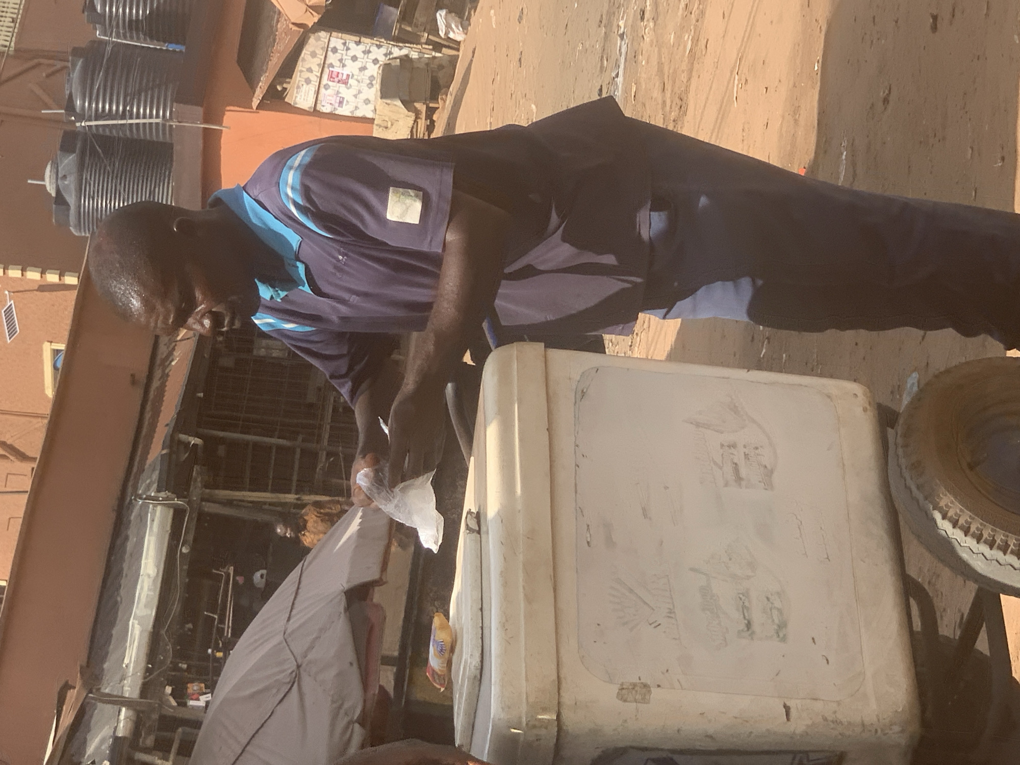 The image captures a street vending scene in Onitsha, Nigeria, on a bright, sunny day. A dark-skinned man, appearing middle-aged, is the central figure. He is dressed in a dark blue polo shirt with lighter blue collar and shoulder accents, and dark blue trousers. A light-colored, square patch is visible on his left chest pocket.
The man stands behind a large, white, rectangular cooler or ice chest. He is engaged in an activity involving the cooler's contents, holding a small, clear plastic bag in his hands, likely containing a product for sale. A small yellow and white packet is tucked into the side of the cooler, and the cooler's lid has faint, etched-in drawings resembling simple house shapes.
The ground beneath is unpaved dirt or sand. In the background, several large, black plastic water storage tanks are visible, some stacked. A small solar panel is attached to a building wall further back. To the right of the cooler, a section of a vehicle tire with a metal rim is visible, suggesting the cooler is part of a mobile cart or stall setup. Other structures, including what appears to be a makeshift stall with a grey fabric awning, are visible in the background, along with a patterned building wall. The bright sunlight casts distinct shadows, indicating it is daytime. The man's expression is neutral and focused on his task. No discernible text is clearly legible on any objects or signs.
