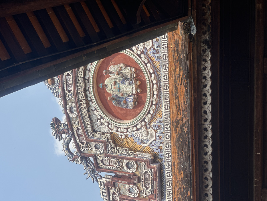 The image displays a highly ornate section of a traditional Vietnamese building's rooftop, set against a light blue sky with minimal cloud cover. The structure, located in Phú Xuân Ward, Hue City, Vietnam, features an extensively decorated gable. Central to this gable is a circular, terracotta-red medallion containing three relief figures. The central figure is a man wearing a tall, patterned hat and lighter-colored robes, flanked by two other men in darker, patterned robes, all standing front-facing.
Surrounding the central medallion and extending across the gable are intricate mosaic patterns composed of white, blue, and brown ceramic pieces, forming geometric designs and stylized floral motifs. Visible at the upper edges of the gable are sections of two stylized dragon sculptures. Below the elaborate gable, rows of weathered orange and reddish-brown terracotta roof tiles are present. To the right of the frame, a dark wooden architectural element, possibly an eave or part of an adjacent roof, diagonally descends into view. The scene is illuminated by bright daylight.