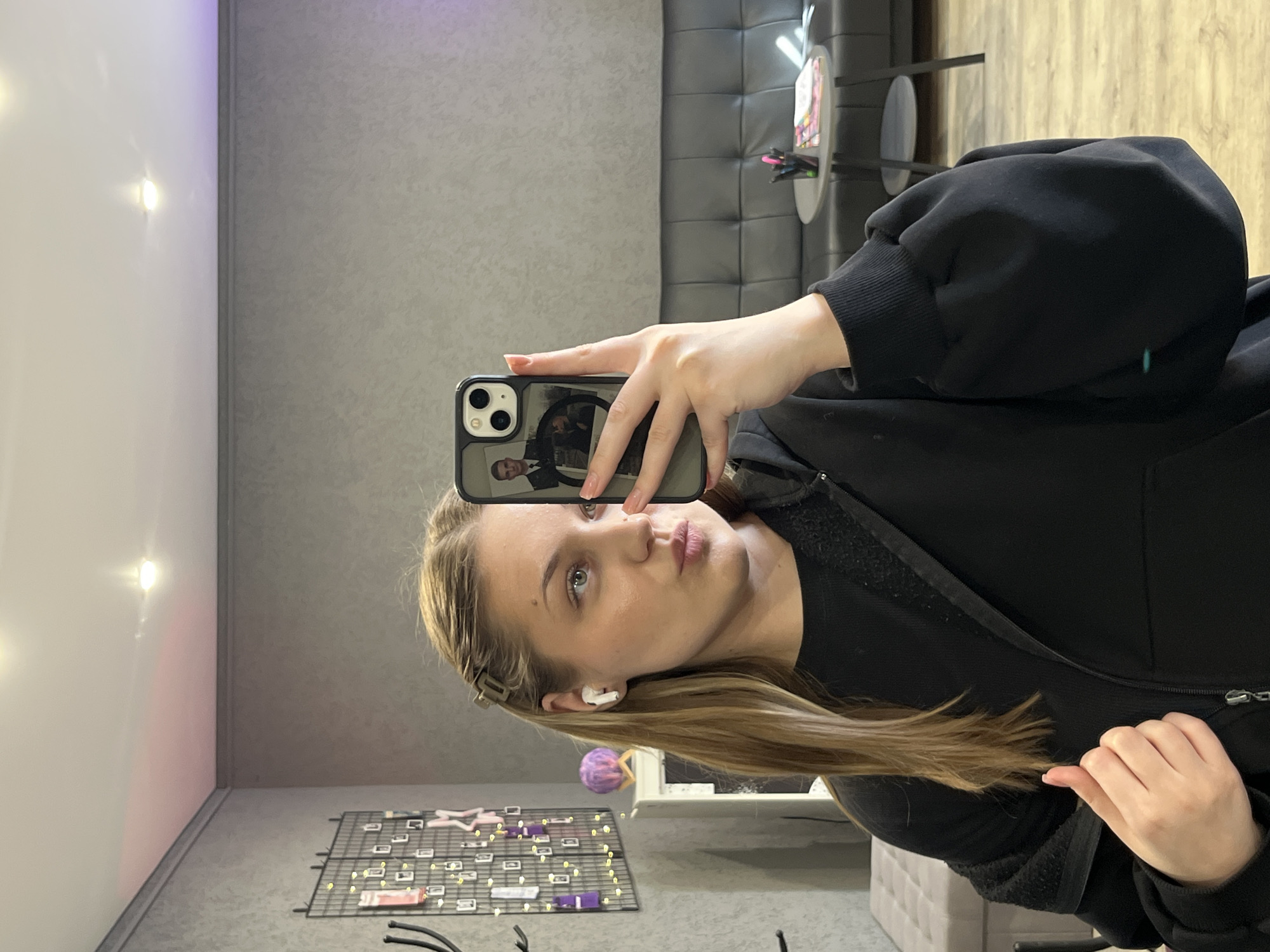 A woman takes a selfie in a room with light grey walls and flooring. She holds an iPhone with a reflective case, showing the reflection of a man in a suit. She is fair-skinned with blonde hair, wearing a black zip-up jacket and has an earbud in her left ear. A decorative metal grid with various items hanging on it sits on the lower left, and a cushioned grey bench is visible in the background on the right side. There's a padded grey wall behind a small circular table with pens and paper. A glimpse of a window with a purple object is seen on the bottom. The location might be a beauty salon or a similar commercial space, however, it can't be determined if the room is located in Rostov-on-Don, Russia, without further data.