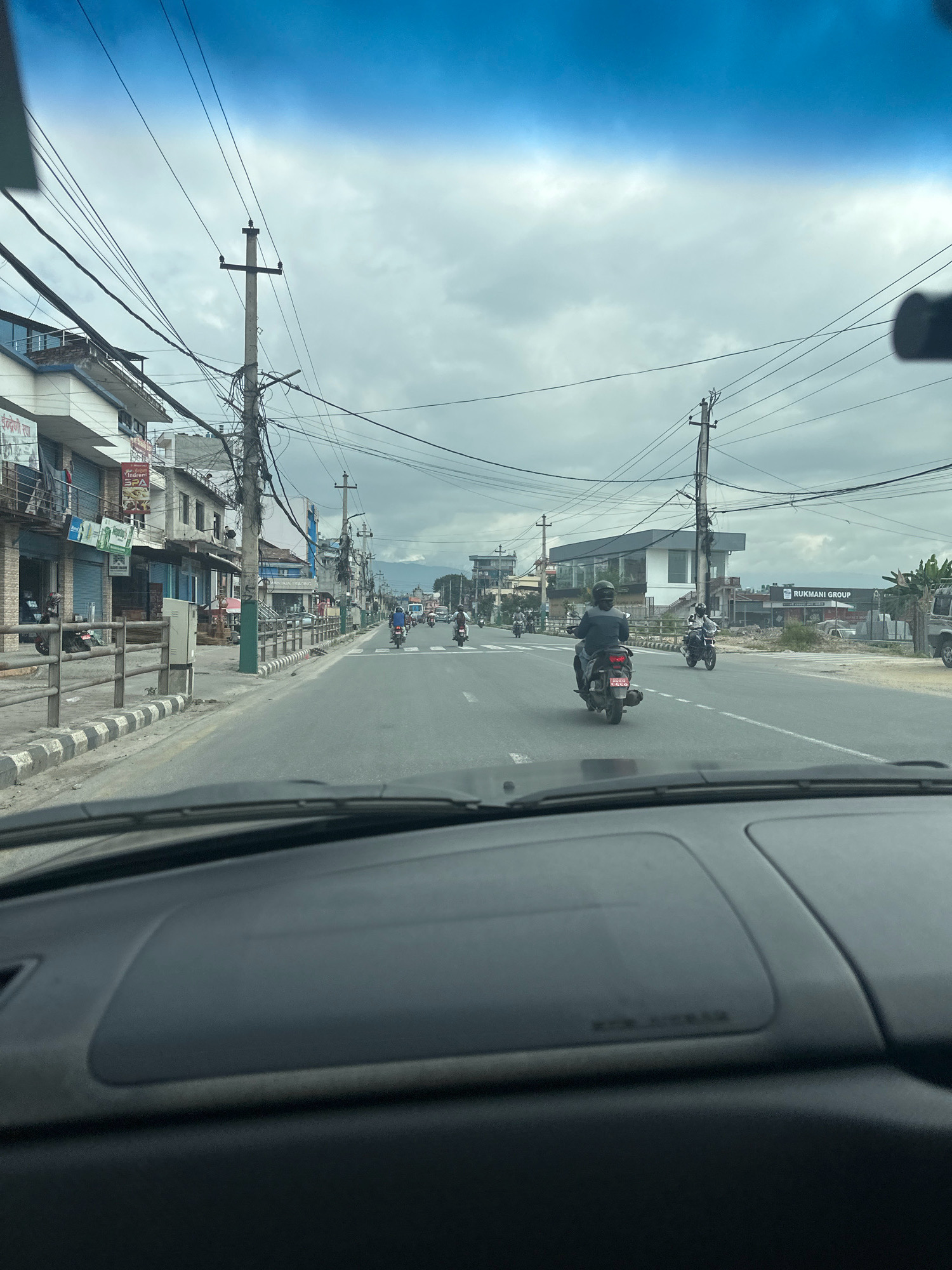 The image captures a view from inside a vehicle, looking out at a street scene in Kathmandu, Nepal. The sky is overcast with heavy clouds. The road is paved and appears to be a main thoroughfare, with multiple lanes of traffic. Several motorcycles are visible, with riders wearing helmets. On the left side of the road, there are buildings that appear to be shops or businesses, with signs in what seems to be Nepali script. There are numerous utility poles and a complex network of electrical wires overhead. A metal railing with a striped pattern separates the sidewalk from the road in some sections. On the right side of the road, there are more buildings and a sign for "RUKMANI GROUP". The foreground shows the interior of the vehicle's dashboard.