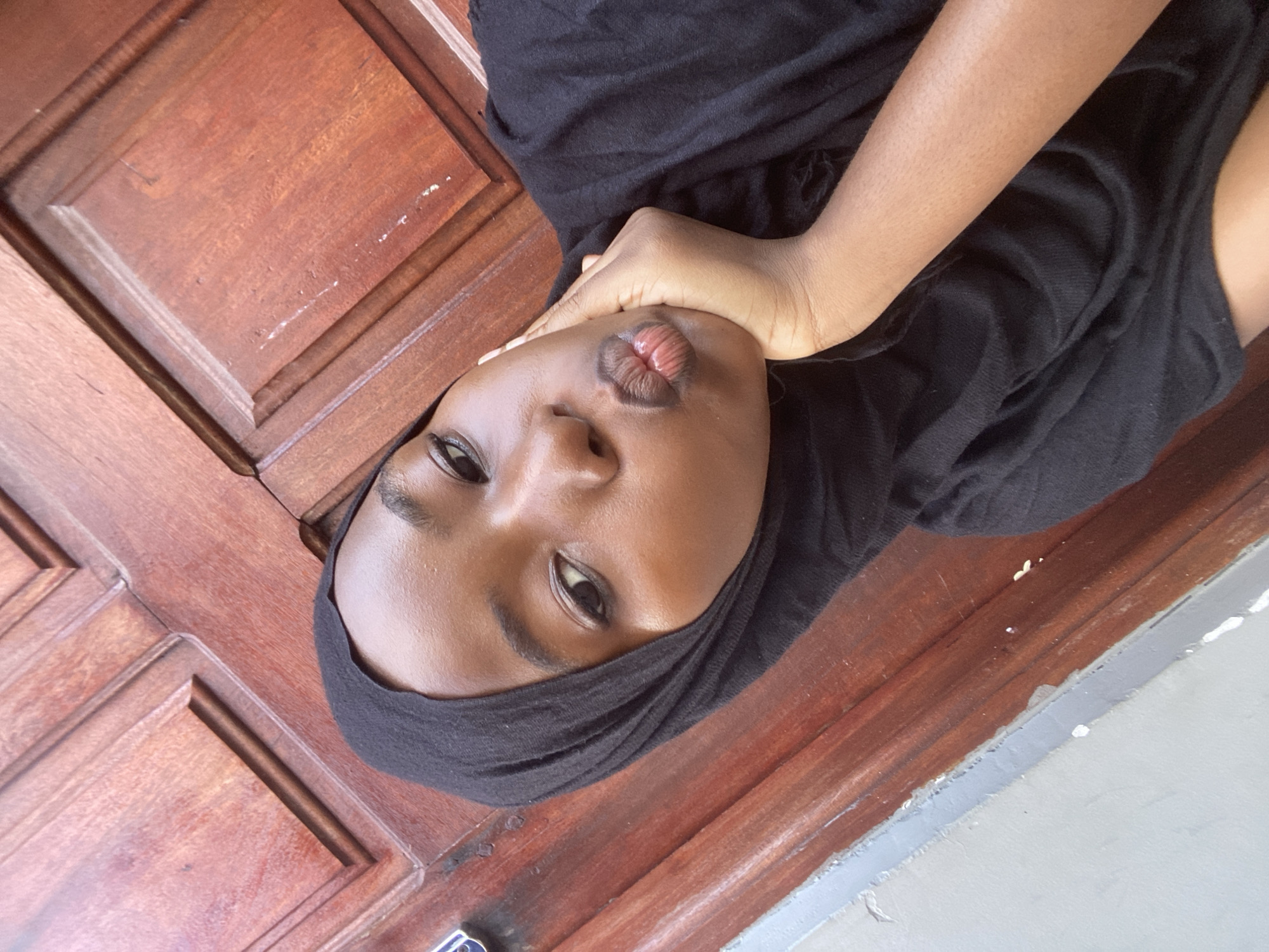 A close-up, angled photograph captures an individual, likely female, with a dark complexion, wearing a black hijab and a black garment. The person's head is tilted slightly, and her lips are pursed in a "duck face" or pouting expression. Her right hand is positioned under her chin, with fingers gently curled. Her eyes, with visible eyeliner, are directed towards the camera. The background consists primarily of a reddish-brown wooden surface, appearing to be a paneled door or wall, with characteristic raised rectangular sections and visible wood grain. Scattered light-colored specks are present on the wooden surface. In the upper left portion of the background, a section of a light gray wall is visible, separated from the wooden surface by a narrow strip of darker gray trim. The specific location within Dar es-Salaam, Tanzania, is not identifiable from the visual content of the image.