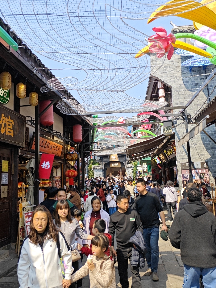 A bustling street scene in a Chinese town. The narrow cobblestone street is packed with people walking in both directions. Traditional Chinese architecture flanks both sides, with shops and restaurants adorned with red lanterns and colorful signage. Overhead, a decorative net of wire and fabric is stretched across the street, with occasional floral and umbrella-like decorations. The sky is bright blue, and the overall atmosphere is vibrant and lively.