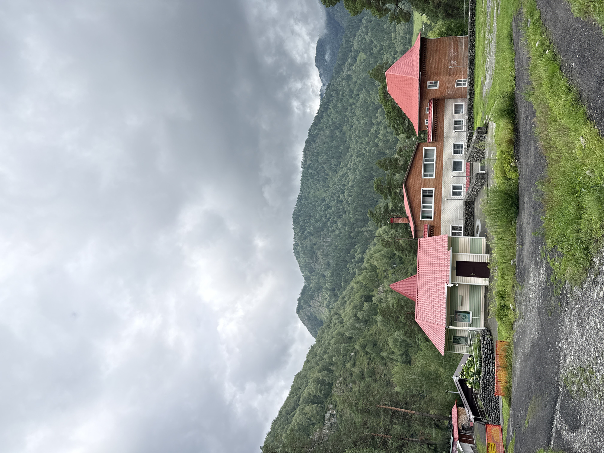 In Chemal, Russia, a cluster of buildings with red roofs stands against a backdrop of lush, green mountains under a cloudy sky. The main building is a two-story brick structure with numerous windows. Adjacent to it is a smaller, mint-green and white building with a distinct conical red roof. Further to the left, a dark-roofed structure with a stone wall is visible, and a low, red-tiled roof building is also present. The foreground shows a gravelly, dark-colored path and patches of green grass. The overall scene depicts a serene, possibly rural or resort-like setting.