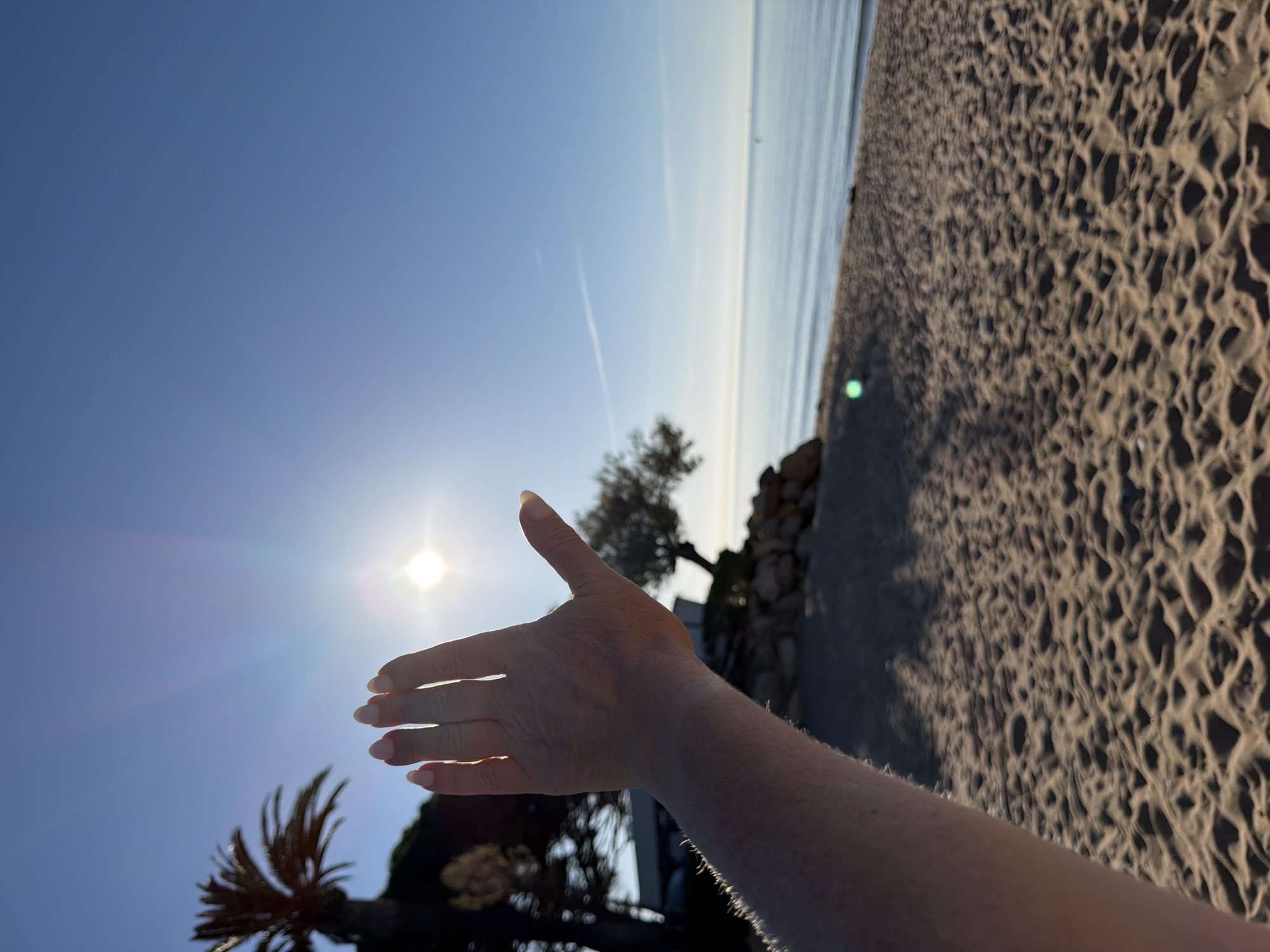 The scene is a sunny beach at Mont-roig del Camp, Spain. A person's arm and hand are extended towards the sun, partially obscuring it. The foreground shows rippled sand. In the mid-ground, a small tree and possibly a rock formation are visible. The background consists of the sea and the clear blue sky. A contrail from an airplane is faintly visible in the sky. A palm tree is in the left corner.