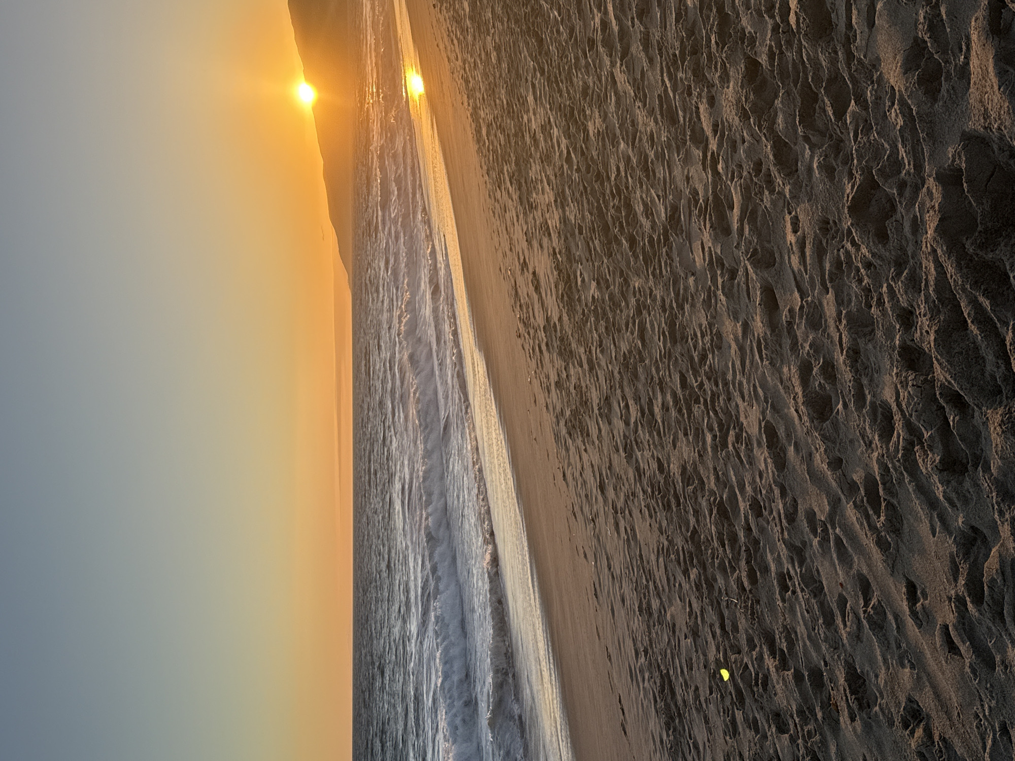 An ocean sunset scene at a beach in Malibu, California. The sun is low on the horizon, casting a warm orange glow across the sky and reflecting on the water. Gentle waves are rolling onto a sandy shore. The sand in the foreground is textured with footprints and appears to have some small debris on it. In the distance, a hilly coastline is silhouetted against the sunset. The sky transitions from a light blue at the top to a warm orange near the horizon.