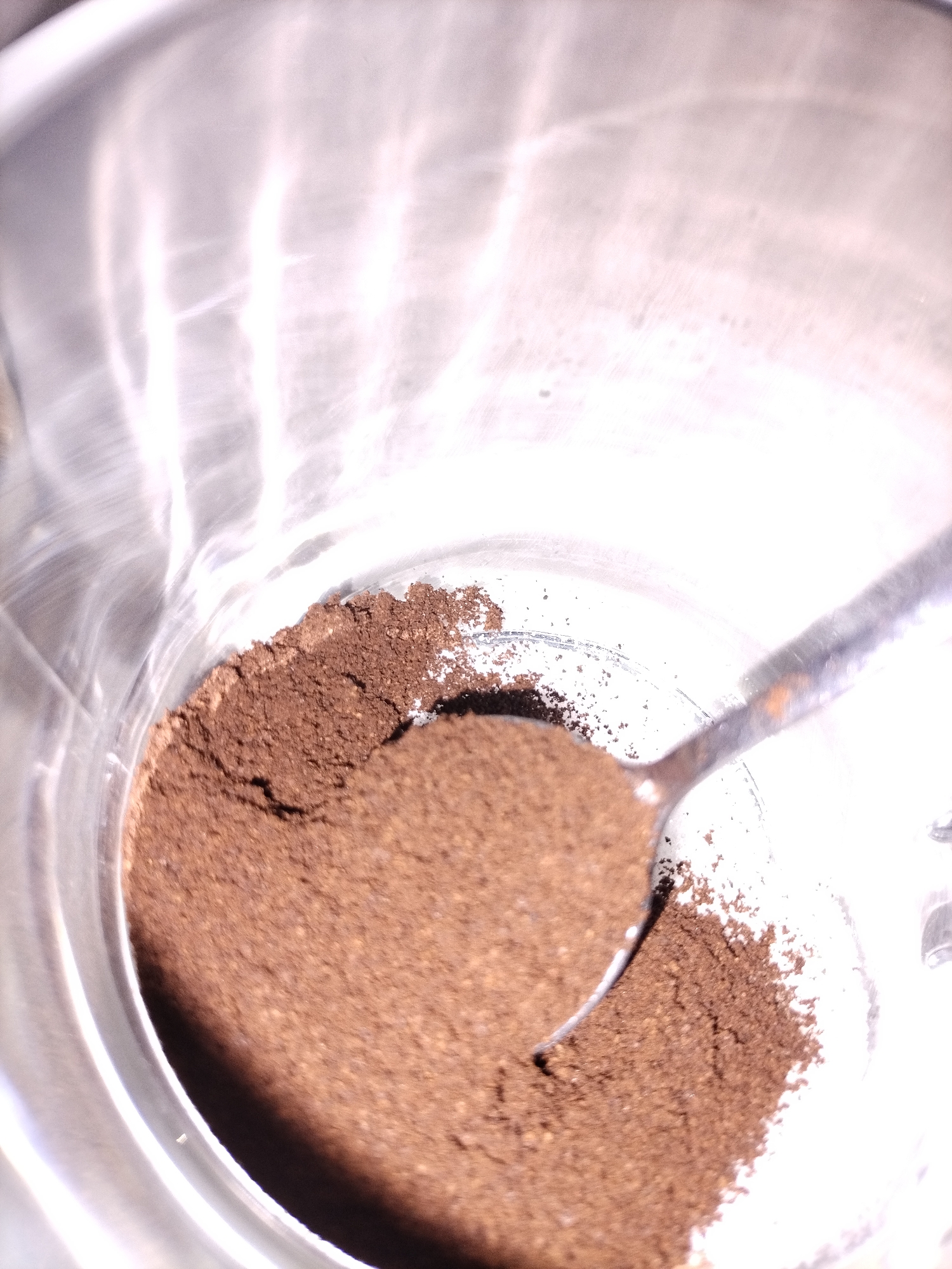 A close-up, top-down shot shows a small pile of dark brown coffee grounds in a clear glass container. A metal spoon rests in the grounds, with its bowl partially filled and some grounds scattered around it. The glass container has bright reflections, suggesting it's a shiny metallic surface or illuminated by a bright light source. The background is out of focus, with streaks of light and shadow. No people or specific location details are visible in the image.
