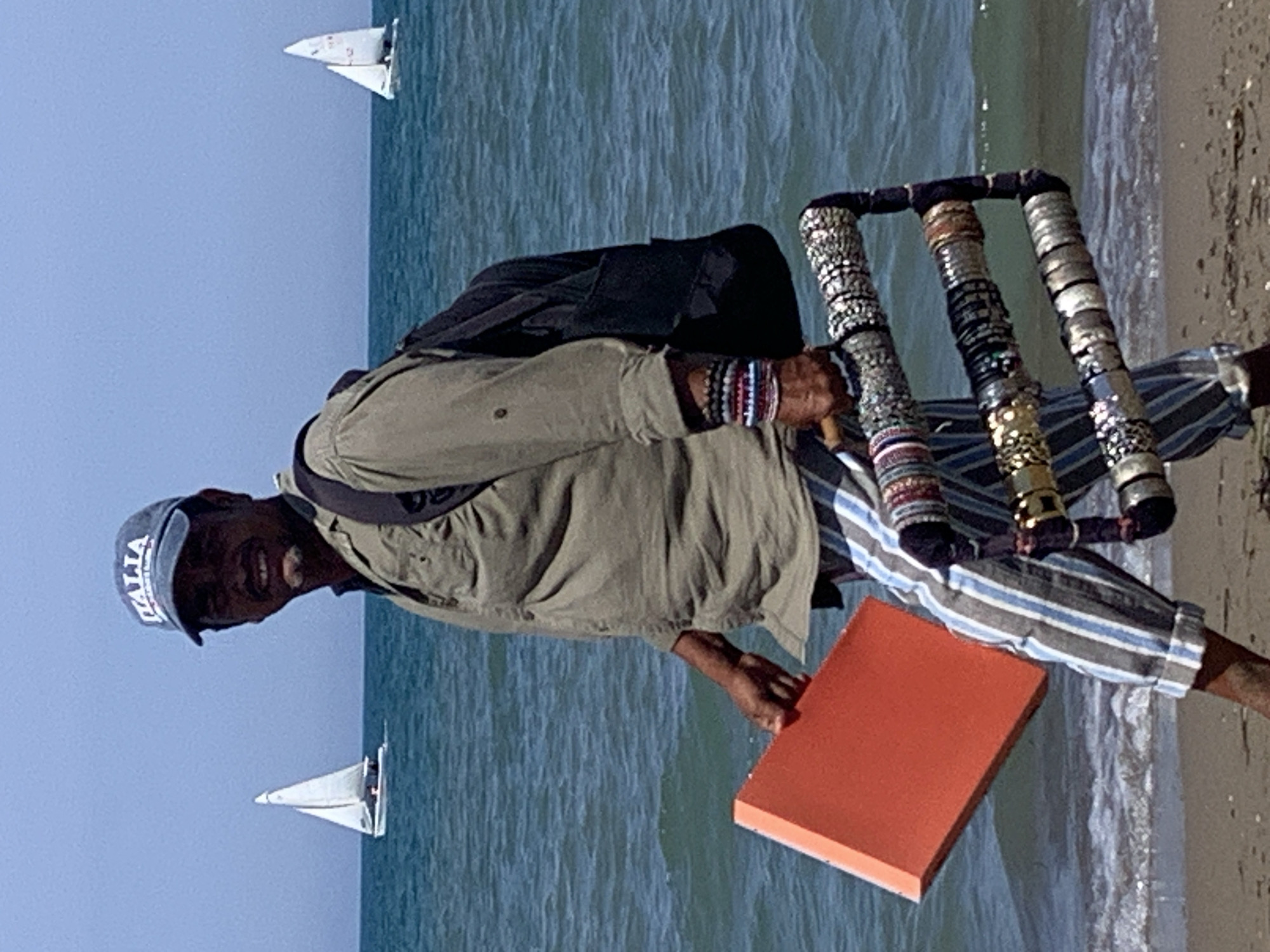 A man wearing a blue baseball cap with "ITALIA" printed on it and a light olive-green shirt, striped pants, and a black backpack walks along the beach near the water. He holds an orange, rectangular object in his left hand and a display stand with multiple bracelets in his right. The sea is blue with waves, and two sailboats are visible in the background. The scene appears to be a sunny day.