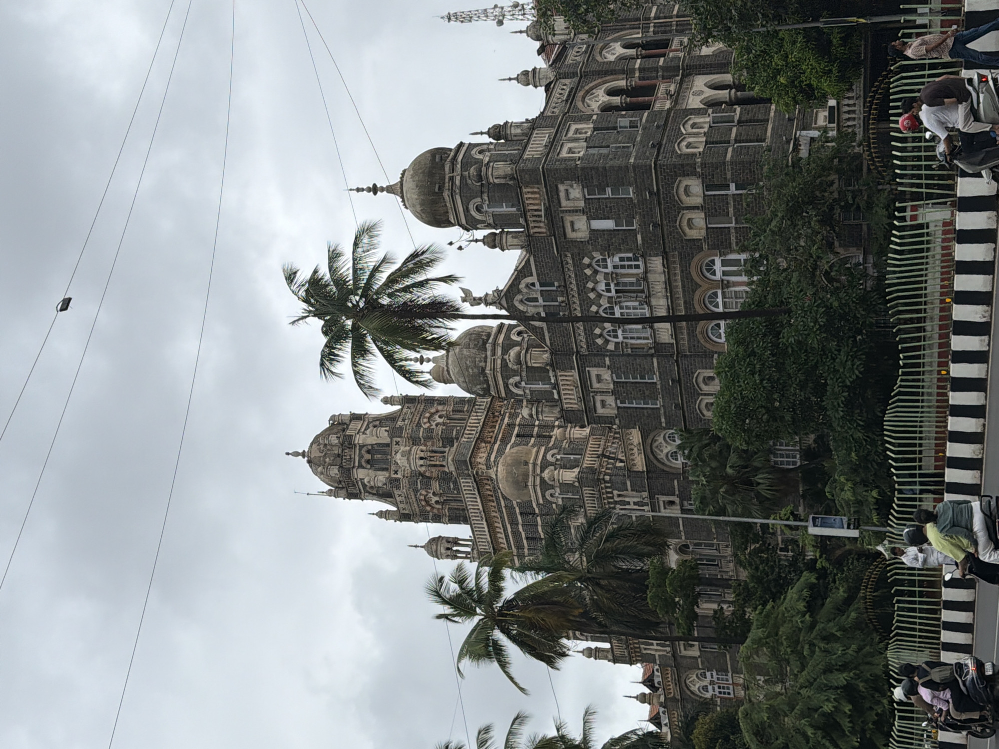 The image depicts the Chhatrapati Shivaji Maharaj Terminus (CSMT) in Mumbai, India, a UNESCO World Heritage Site and a historic railway station. The grand Victorian Gothic Revival architecture of the building is prominent, characterized by its intricate stonework, domes, and tall spires, set against an overcast sky. Several palm trees are visible in the foreground, framing the majestic structure. A busy road runs along the front of the station, with traffic including motorcycles and cars. People are seen riding on motorcycles and walking on the sidewalk. The scene captures a typical day at this iconic Mumbai landmark, blending historical grandeur with the city's vibrant street life.