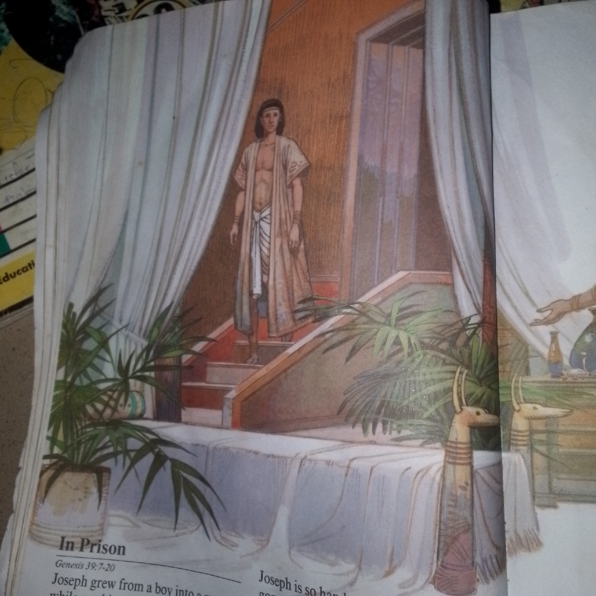 The image depicts an illustration of a scene from the book of Genesis, specifically illustrating Joseph in prison. The setting appears to be an interior space, likely a room in ancient Egypt, suggested by the style of dress and the decorative elements. A young man, presumably Joseph, stands on a short set of stairs, dressed in a simple, draped garment that exposes his chest and torso, with a white cloth tied around his waist. He has dark hair and a serious expression. He is positioned near a doorway and in front of flowing white curtains. To the left, a potted plant with long, green fronds is visible. The stairs lead up to a landing where a table is covered with a white cloth. Two decorative objects with animal-like heads and striped bodies stand on either side of the table, draped with fabric. In the background, a doorway with vertical panels is visible. The text at the bottom of the image indicates "In Prison" and references Genesis 39:7-20. The overall atmosphere is one of quiet contemplation or waiting, consistent with the title "In Prison." The location is not specified in terms of a city or known geographical area, beyond the implied ancient Egyptian context.