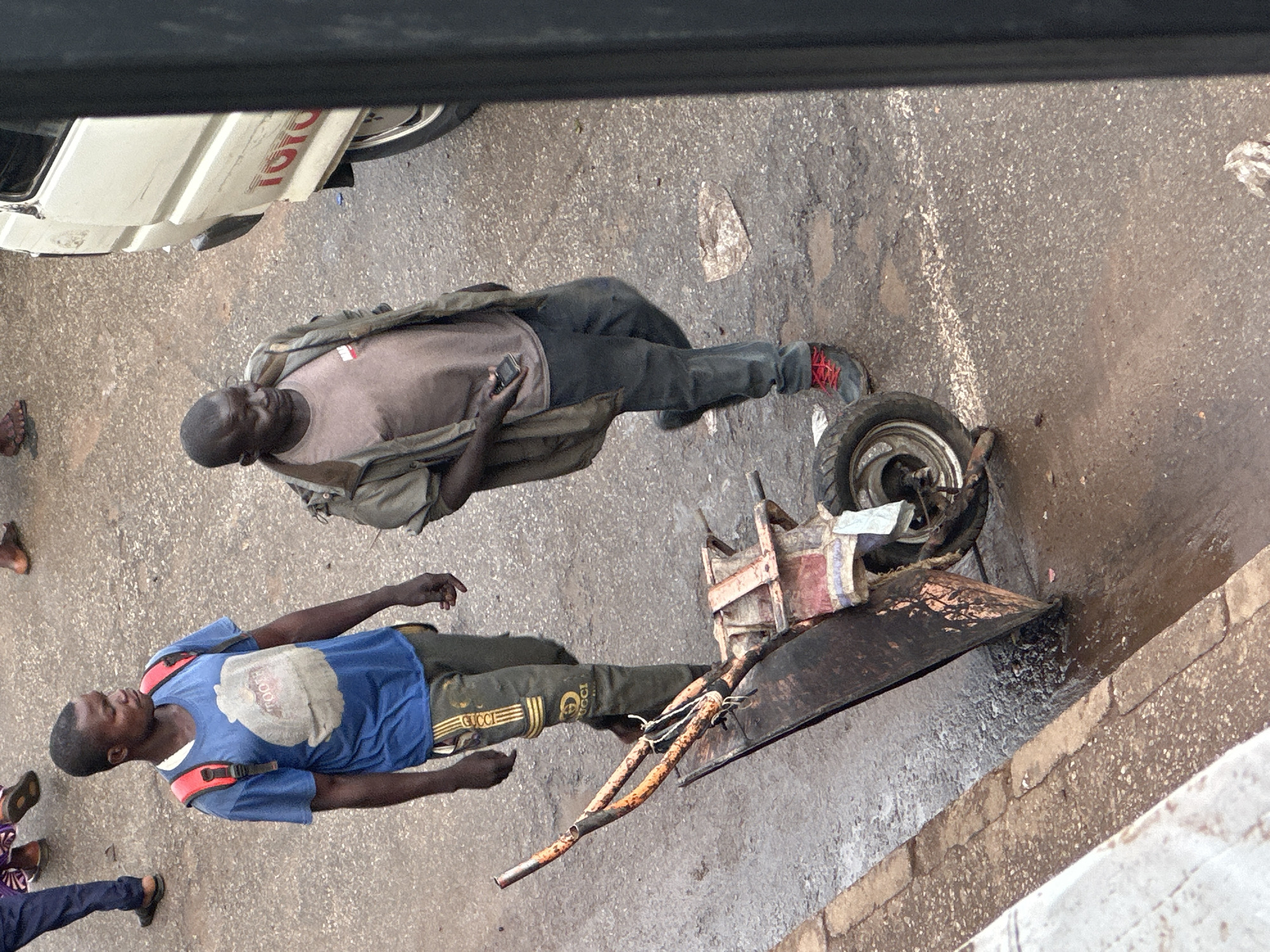 This media file shows two young men and parts of a vehicle on a paved surface, likely a road. One man, wearing a blue t-shirt with a graphic design and dark pants, is lying down on his back, facing upwards. He has a red strap across his chest, suggesting he might be wearing a backpack. The other man, wearing a light brown t-shirt and dark pants, is also lying down, on his side, with his head resting on his arm. He is holding a mobile phone in his hand.
Nearby, a partially disassembled motor scooter is visible, with its wheel, frame, and some metallic parts scattered on the ground. The scooter appears to be old and worn. In the background, the side of a white vehicle with the red "TOYOTA" logo is visible.
The scene appears to be outdoors, possibly in a street or a workshop, given the presence of the vehicle parts and the paved ground. The lighting suggests it is daytime, and there are no strong indicators of specific weather conditions. The overall impression is one of a casual, perhaps slightly disordered, setting. The facial expressions of the men are not clearly discernible, but their relaxed postures might indicate a moment of rest or conversation. The presence of the scooter parts could suggest that they are working on it or have recently finished a task. The context provided indicates this is Bukuru, Nigeria.
