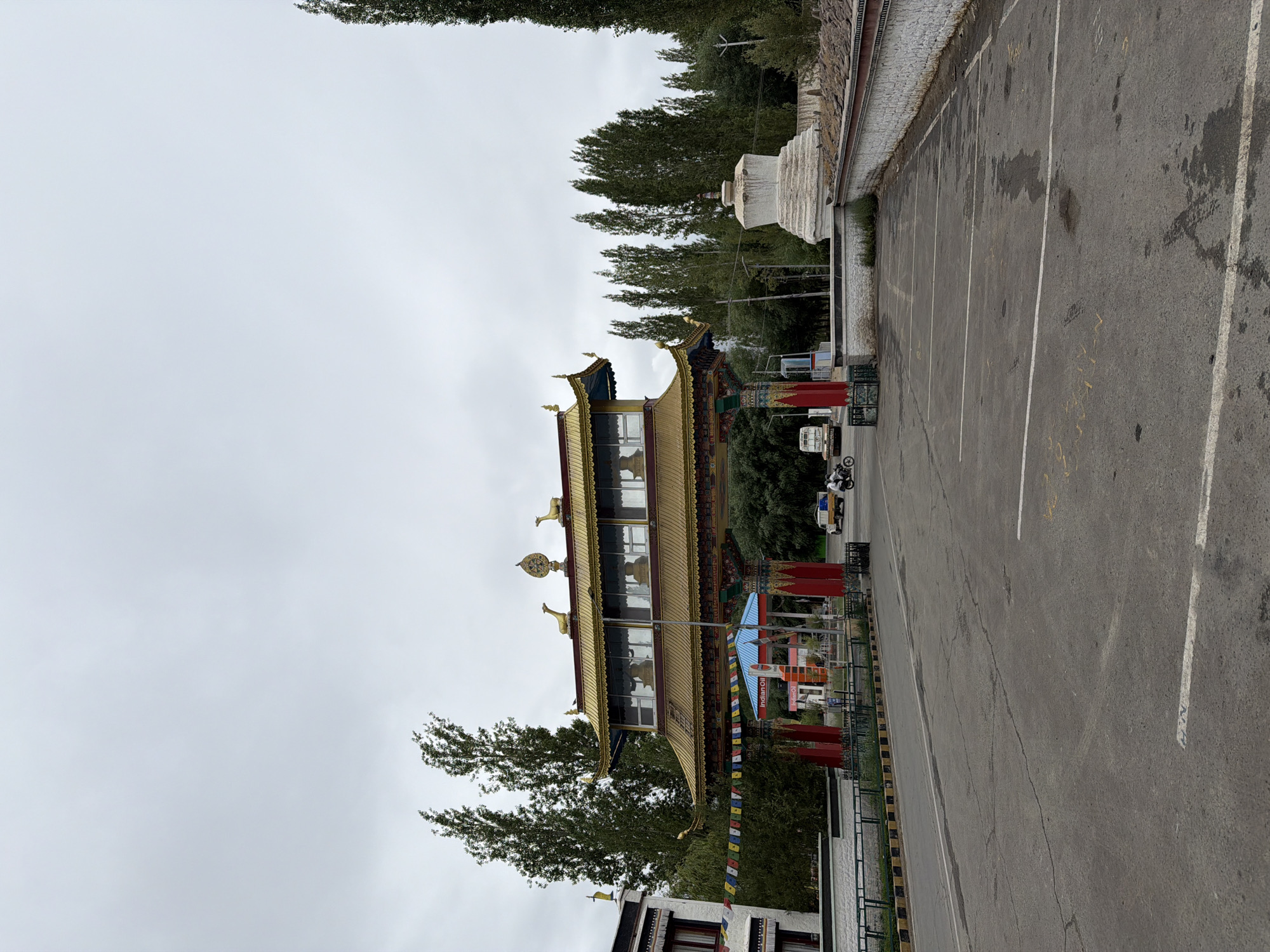 The image depicts a wide-angle, eye-level outdoor shot of a Buddhist monastery gateway in Thiksey, India, under a cloudy sky. The gateway, a prominent feature, is adorned with gold and vibrant colors, featuring multiple tiers and decorative elements, including golden deer statues and a dharma wheel on the apex. Prayer flags are strung across the gateway. To the left of the gateway is a white building with red-trimmed windows. To the right, a white stupa stands prominently.
In the foreground, a paved parking lot with white parking lines is visible. A dark asphalt road runs in front of the gateway. On the road, a white truck and two motorcycles are present. One motorcycle carries a rider and a passenger, while the other appears to have one rider. A blue and red structure, possibly a fuel station, is situated behind the gateway. Tall, slender trees line the background, extending into the distance. The overall environment suggests a religious or cultural site with vehicular access.
