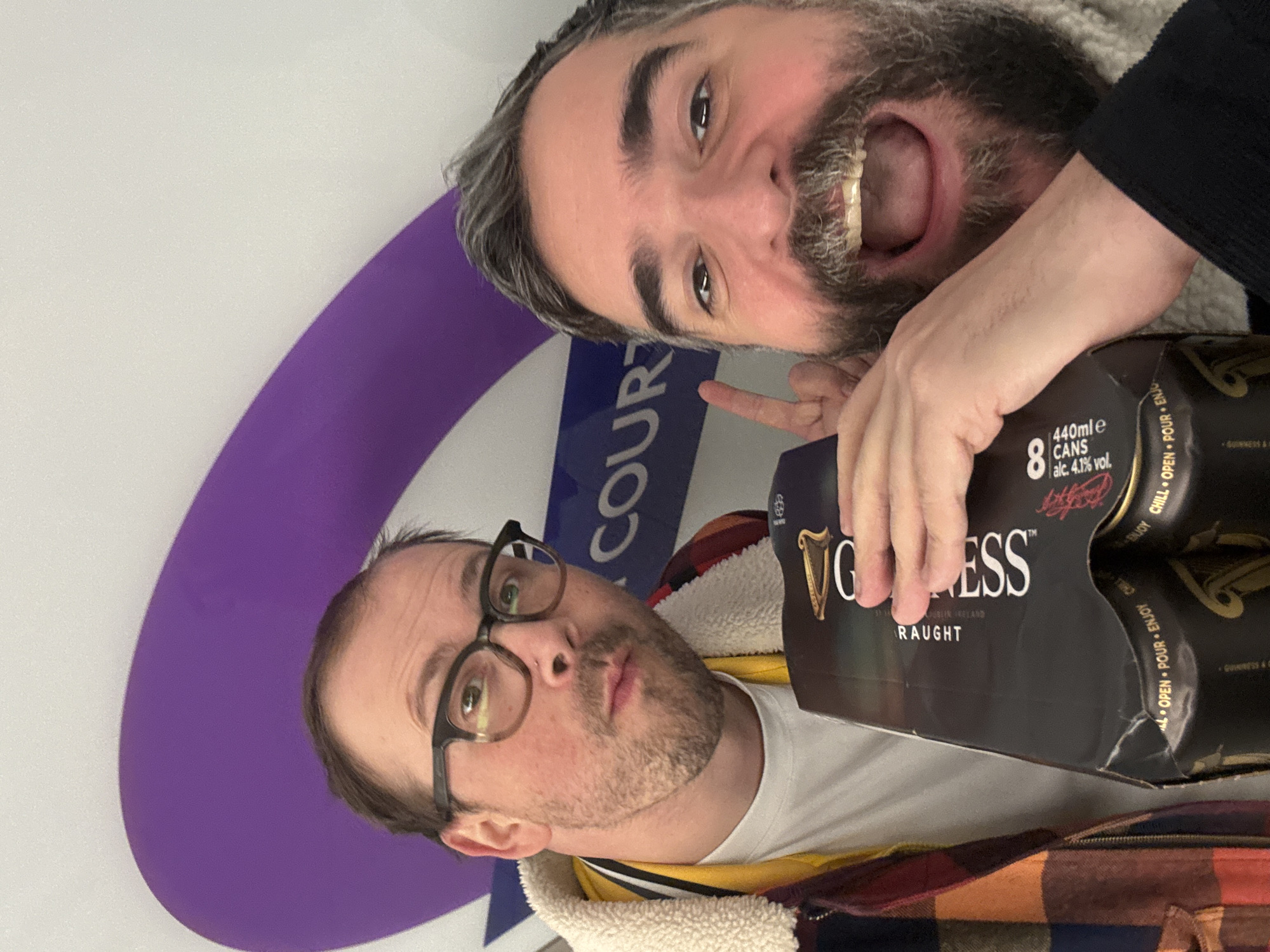 Two men pose for a photo with a pack of Guinness Draught cans. The man in the foreground is wearing glasses and a jacket. The man in the background has a beard and is pointing at the cans of Guinness. In the background, part of a purple and white London Underground logo is visible, specifically the word "COURT", suggesting the photo was taken at a station such as Earl's Court in London, United Kingdom.
