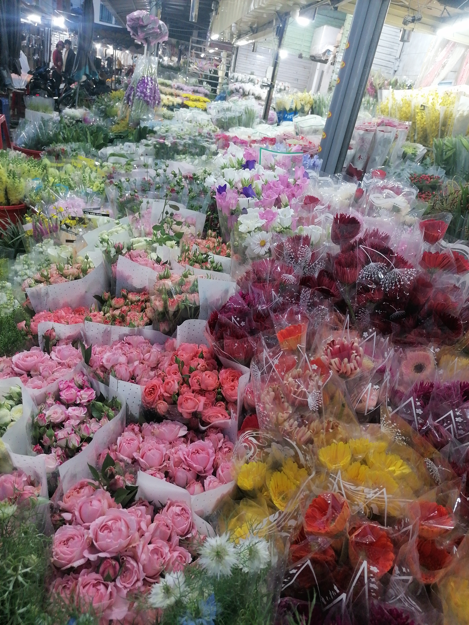 The image depicts a bustling flower market in Da Lat, Vietnam. Numerous bouquets of various flower types, including roses, daisies, and lilies, are arranged in colorful displays. The flowers are predominantly wrapped in clear plastic. The scene is set indoors, possibly under a market stall or shed, with overhead lighting. A metal support structure is visible in the background. A crowd of people is discernible in the background, indicating an active commercial environment.