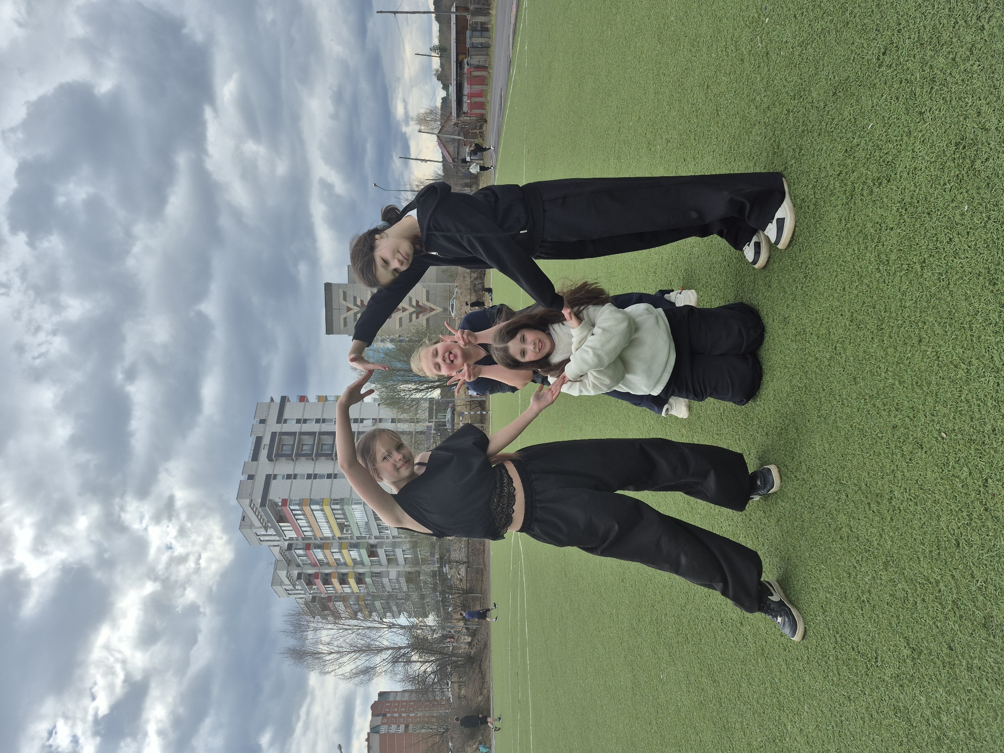 Four young females are playfully posing for a photo on a green artificial turf sports field under an overcast sky. In the background, multi-story residential buildings, some with colorful facades, and bare trees indicate an urban or suburban setting, likely in Chaikovsky, Russia.
On the right side of the frame, a taller girl stands upright, wearing a black tracksuit and black and white sneakers. She smiles at the camera, with her hands resting on the shoulders of two other girls. These two girls are crouching or kneeling in the center; one, with dark hair, wears a light-colored long-sleeved top and dark pants, making a peace sign. The other, with blonde hair, is partially visible and also flashes peace signs with both hands. All three are smiling.
To the left, a fourth girl stands, wearing a black short-sleeved top with a lace hem and black wide-leg trousers, paired with dark sneakers. She holds her arms gracefully above her head, one arm bent, and smiles towards the camera. The overall mood is cheerful and casual, suggesting a friendly gathering or outing. The light is soft and diffused due to the heavy cloud cover, indicating daytime, possibly in cooler weather given the bare trees. No discernible text is visible on clothing or buildings.