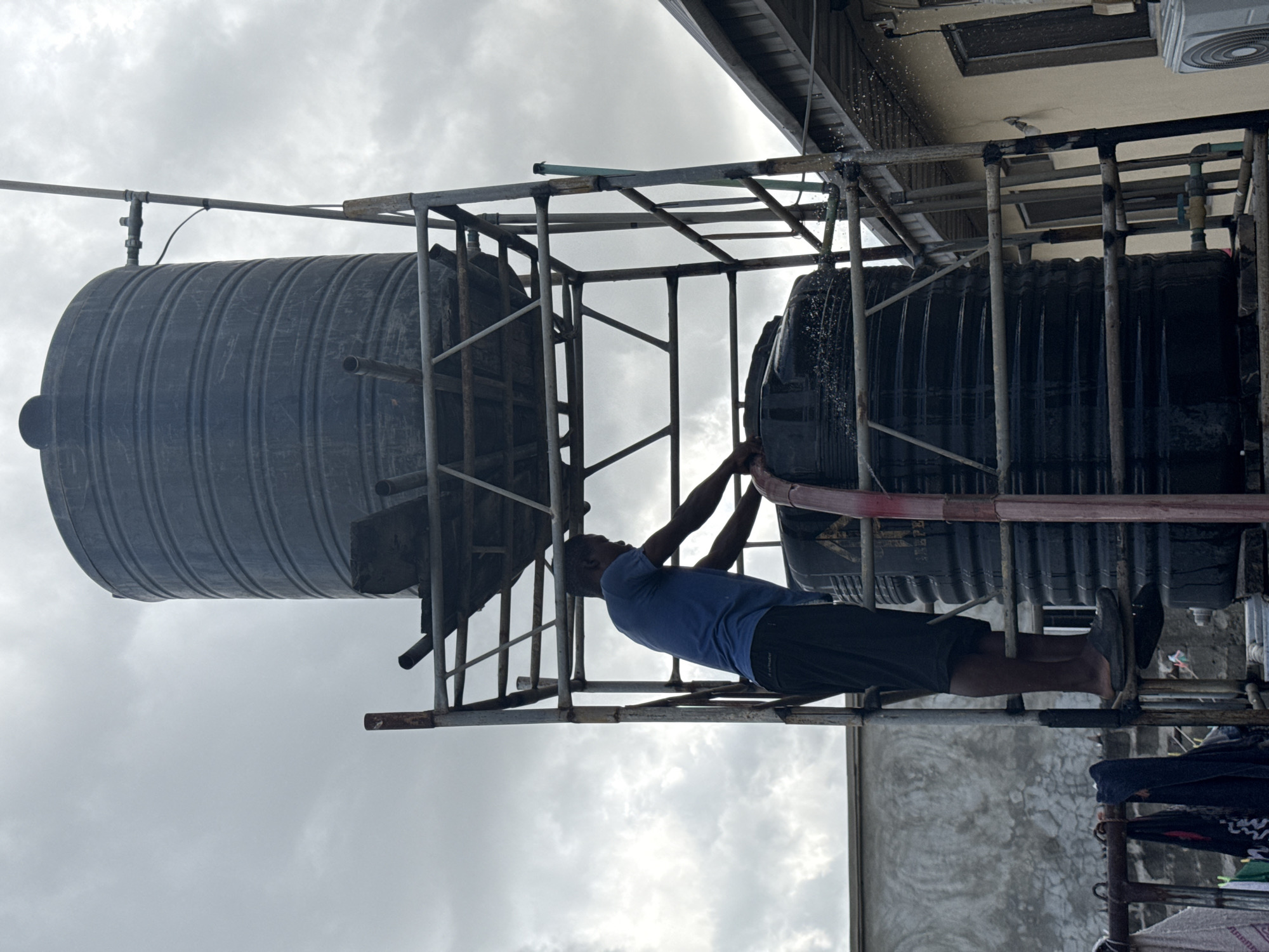 A man is standing on a metal scaffolding structure, reaching towards a pair of large black water tanks elevated on the structure. He is wearing a blue t-shirt and black shorts, with black shoes visible. Water is visibly leaking from one of the tanks. The scaffolding and tanks appear to be situated on a rooftop or elevated platform. The background features a cloudy sky and the top of a building. The location is Ado, Nigeria.