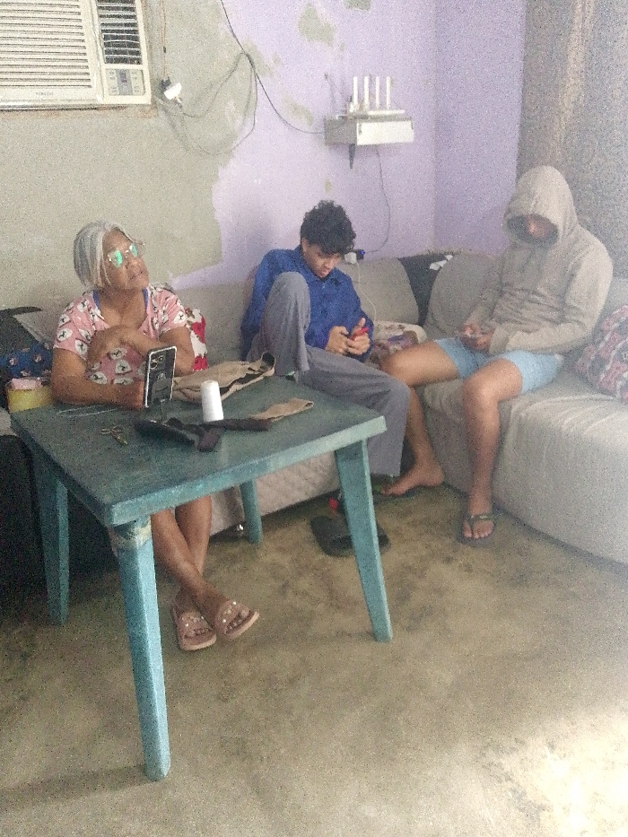 Inside a room with faded walls, three people are present. An elderly woman with glasses sits by a blue table on which rest several items, including a phone, scissors, and a white cup. A young man in a blue shirt and grey pants sits on a couch looking at his phone. Next to him sits another young man wearing a hooded sweatshirt and shorts. They are all looking at phones. A white couch runs along the back wall. An air conditioner sits on the wall above the woman.