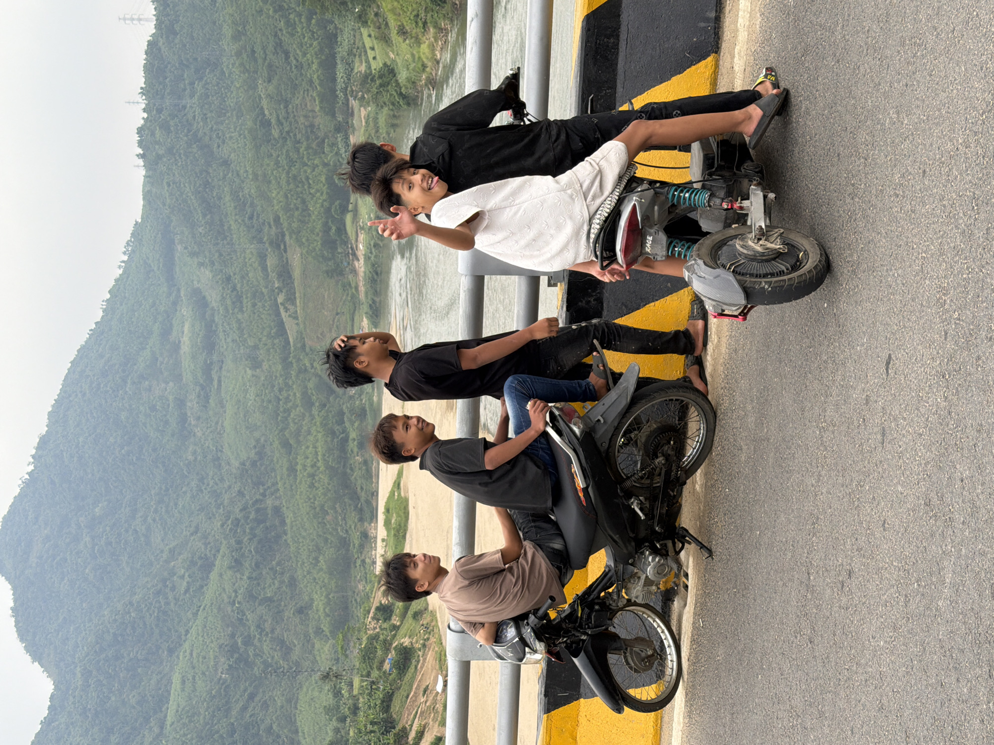 The image shows five young men posing on three motorcycles on a bridge in a rural setting. The background reveals a lush, green, mountainous landscape and a river. The bridge has yellow and black stripes on the edges. One man wears a light brown shirt, another a dark grey shirt with blue jeans, another a black shirt and pants, and another a white shirt. The man on the top of the pile of friends wears a black shirt and pants. Some of the men are wearing sandals. The photo is taken from the side and is angled to emphasize the landscape in the background. The scene appears to be outdoors during the daytime. While the precise location within Đà Nẵng is not discernible from the image, the setting is consistent with the mountainous regions surrounding the city.
